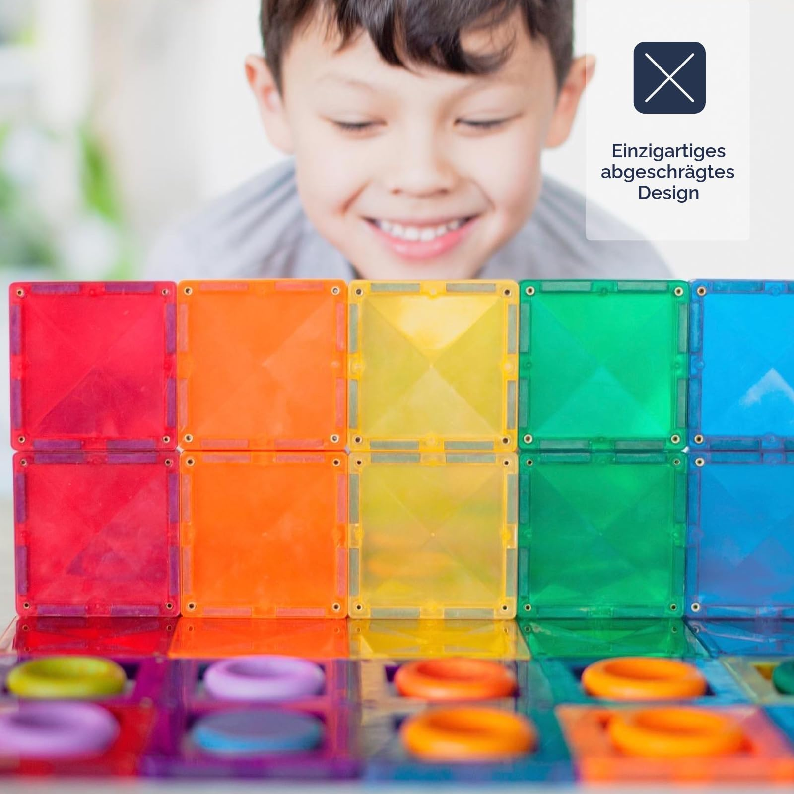 Ein lächelnder Junge baut eine bunte Wand mit magnetischen Quadratblöcken vor sich.