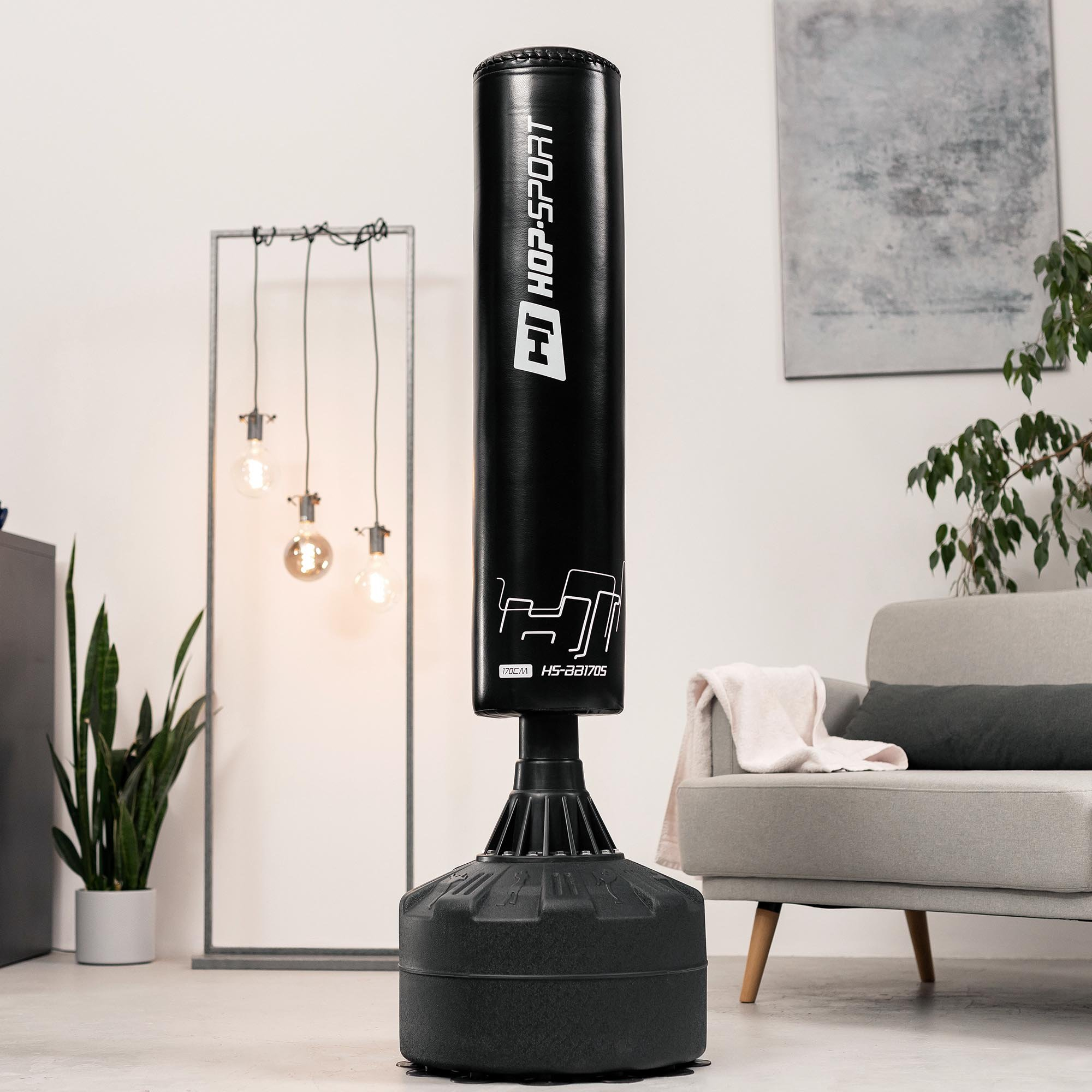 Ein schwarzer Stand-Boxsack von Hop-Sport steht in einem modernen Wohnzimmer mit grauem Sofa und Pflanzen.