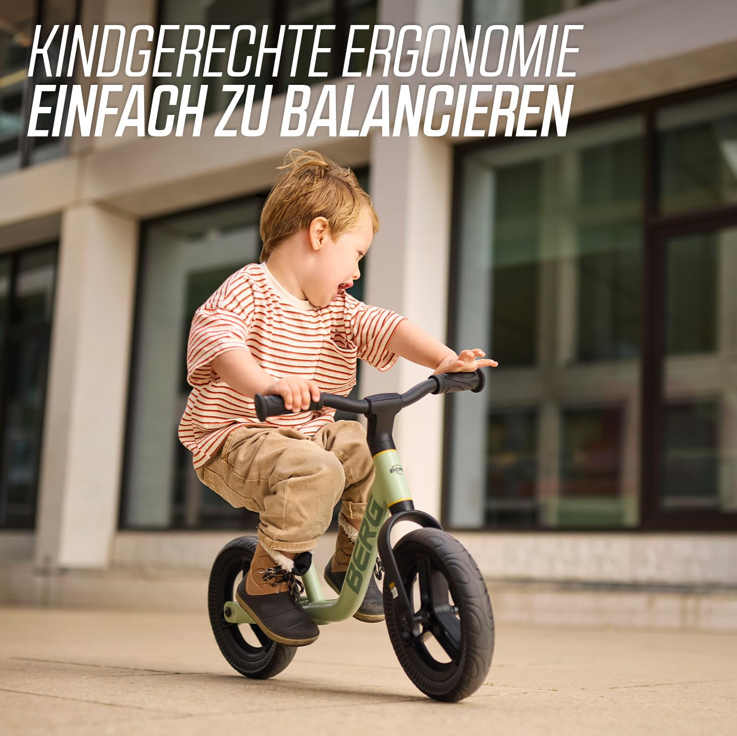 Bambino con maglietta a righe rosse e bianche guida una balance bike verde chiaro davanti a un edificio moderno.