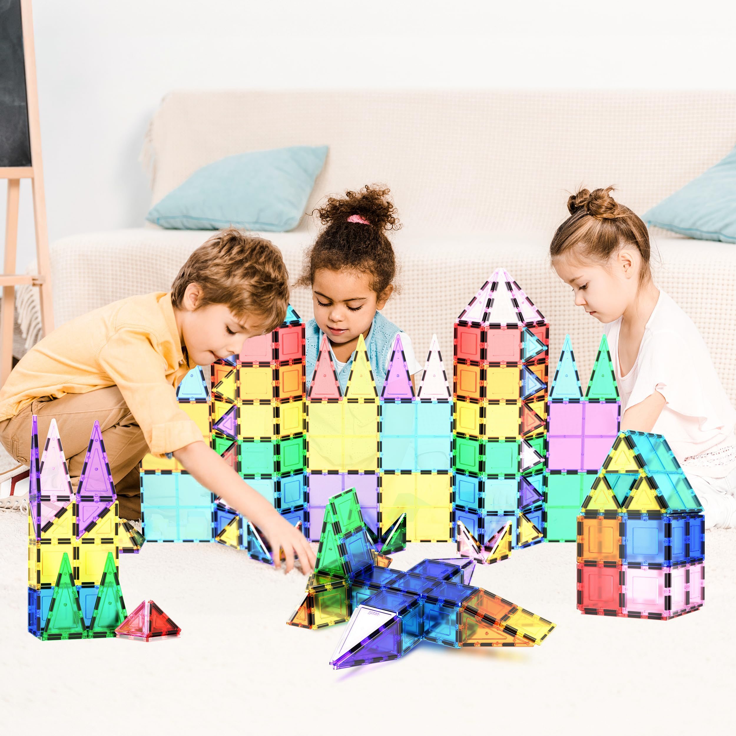 Drei Kinder spielen auf einem weißen Teppich und bauen bunte Konstruktionen aus magnetischen Bausteinen.