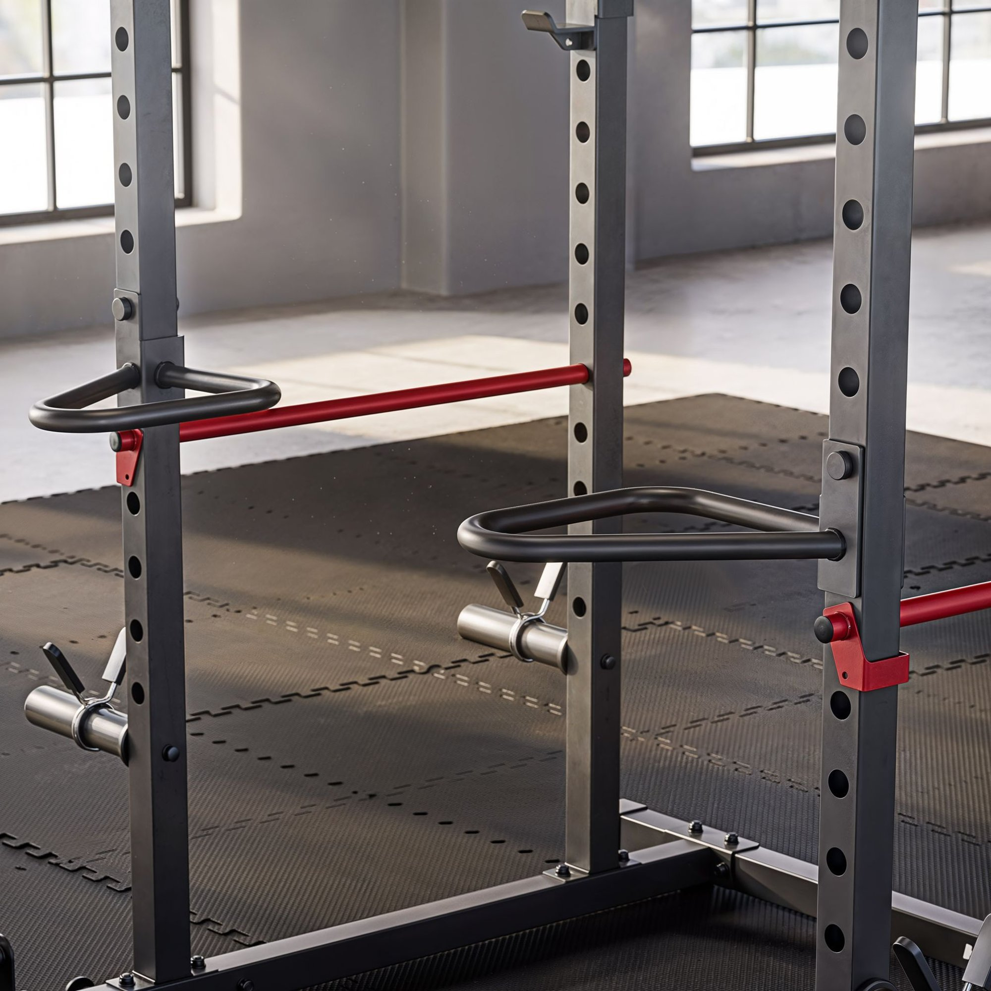 Ein stabiles Fitness-Kniebeugengestell mit roten Akzenten steht auf einer schwarzen Matte in einem Fitnessstudio.