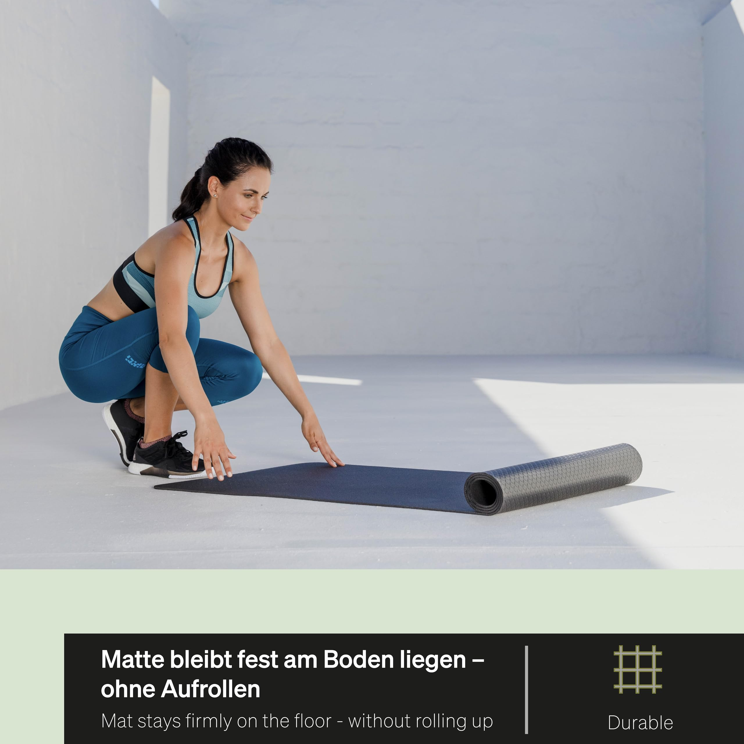 Eine Frau in Sportkleidung rollt auf einem hellen Boden eine schwarze Yogamatte aus.