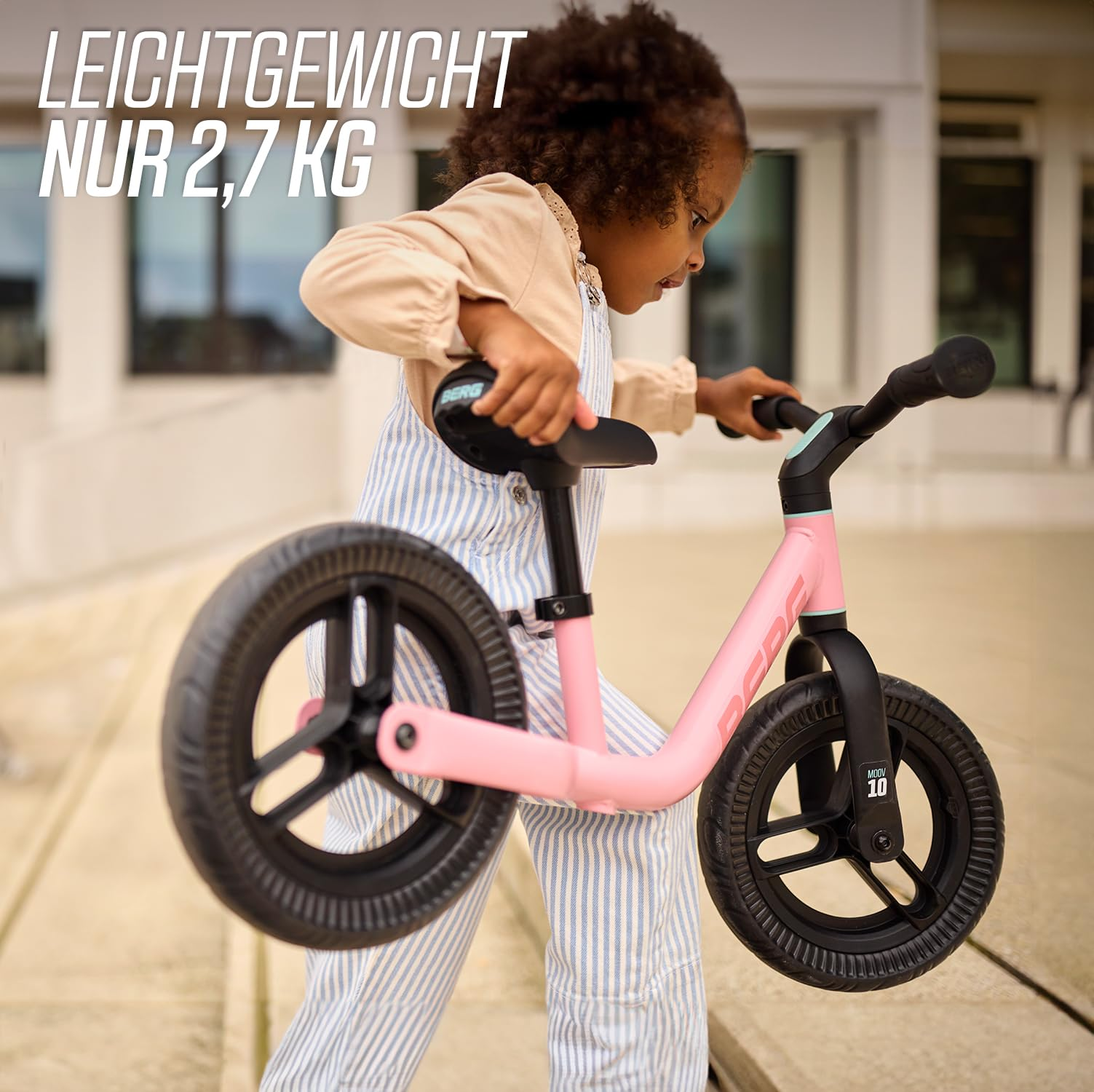 Un bambino con salopette a righe blu e bianche trasporta una leggera bicicletta senza pedali rosa all'aperto.