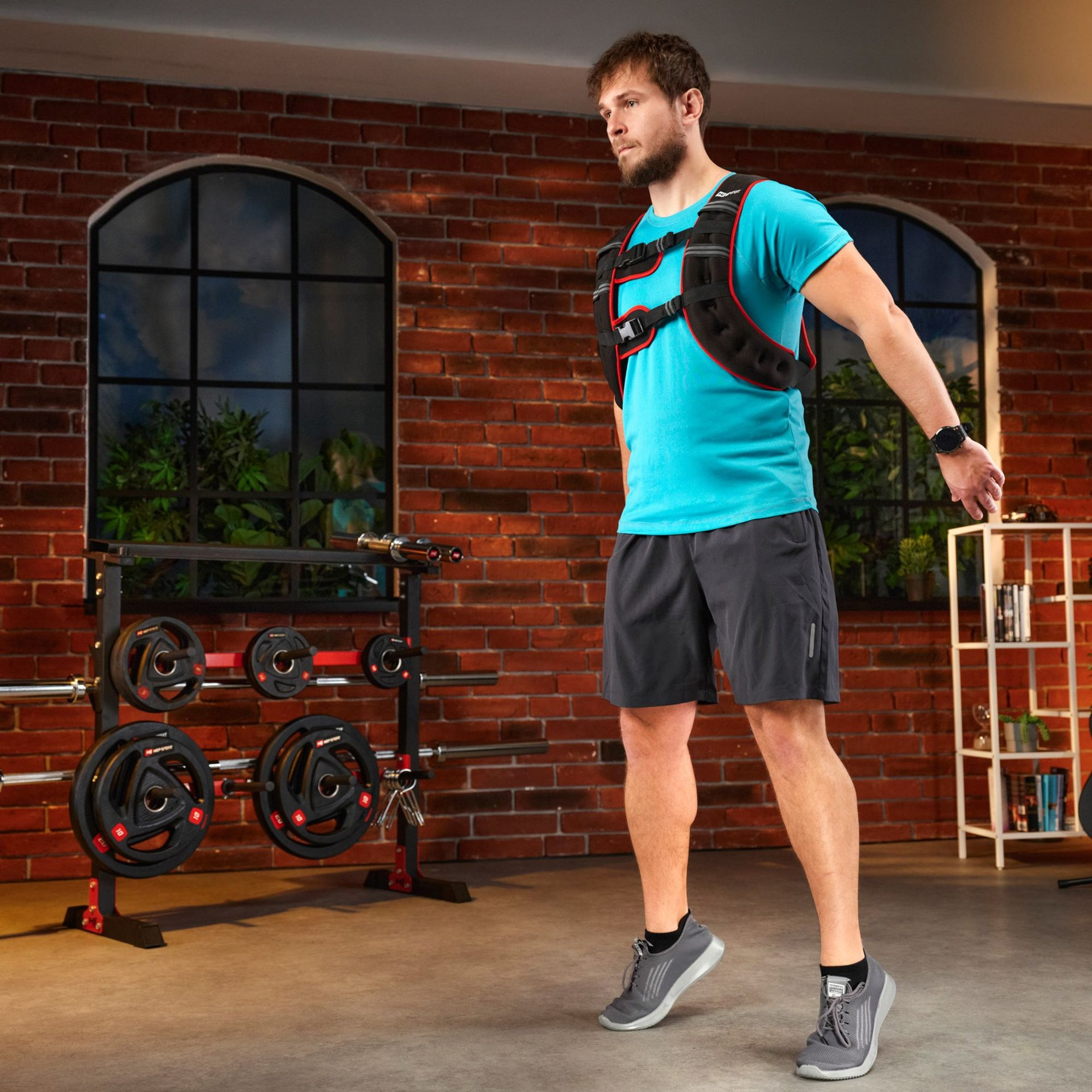Ein Mann mit Gewichtsweste und blauem Shirt springt in einem Fitnessstudio mit Ziegelwand.