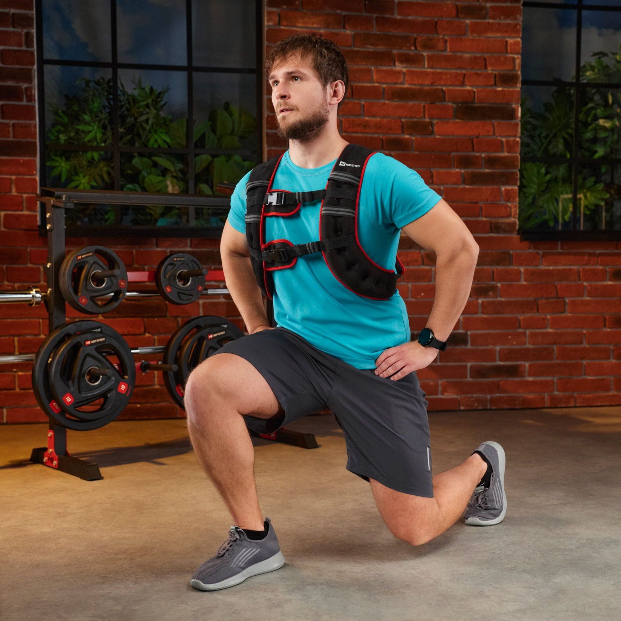 Ein Mann mit blauem Shirt und Gewichtsweste macht Ausfallschritte im Fitnessstudio.