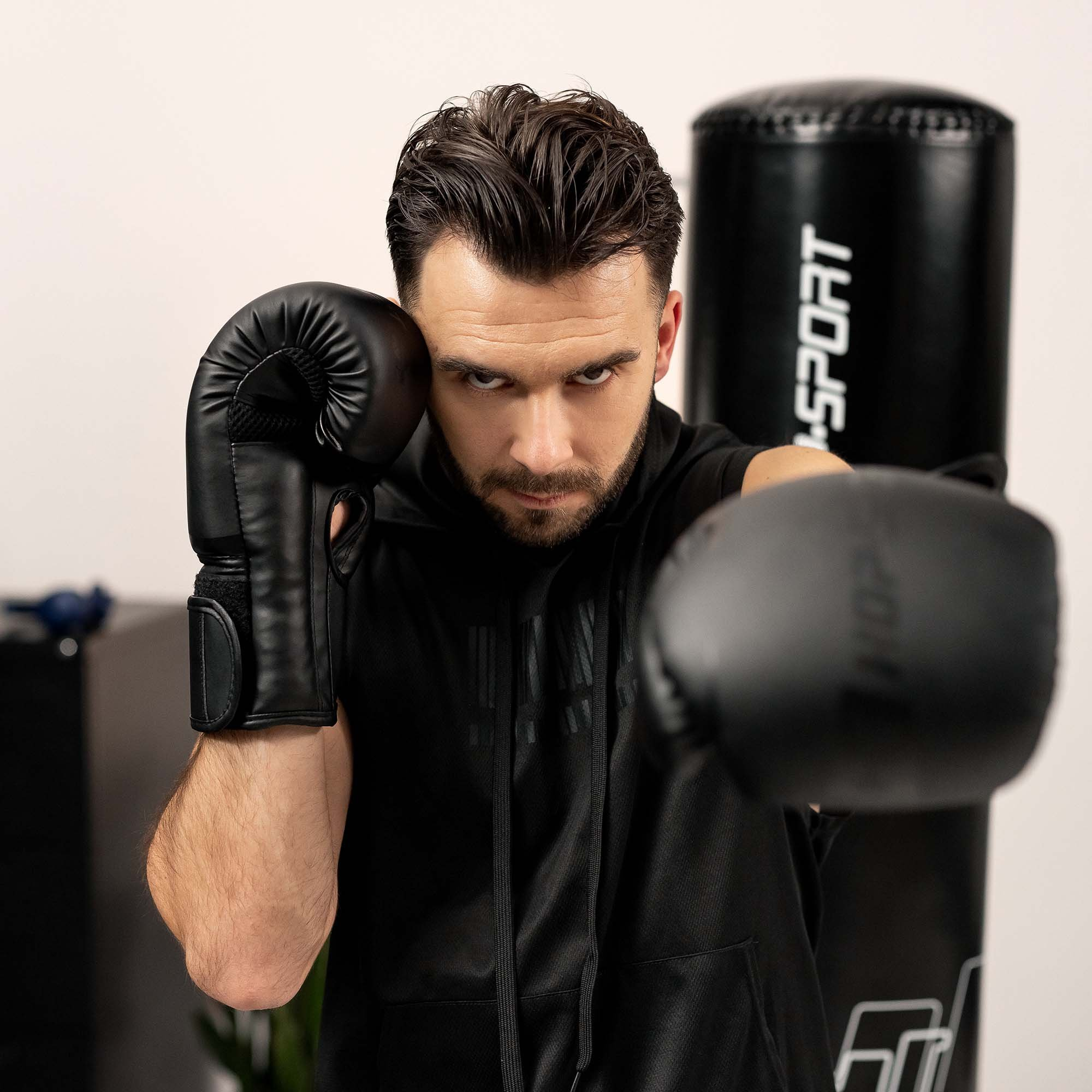 Ein Mann mit schwarzem Oberteil und Handschuhen boxt vor einem Boxsack.