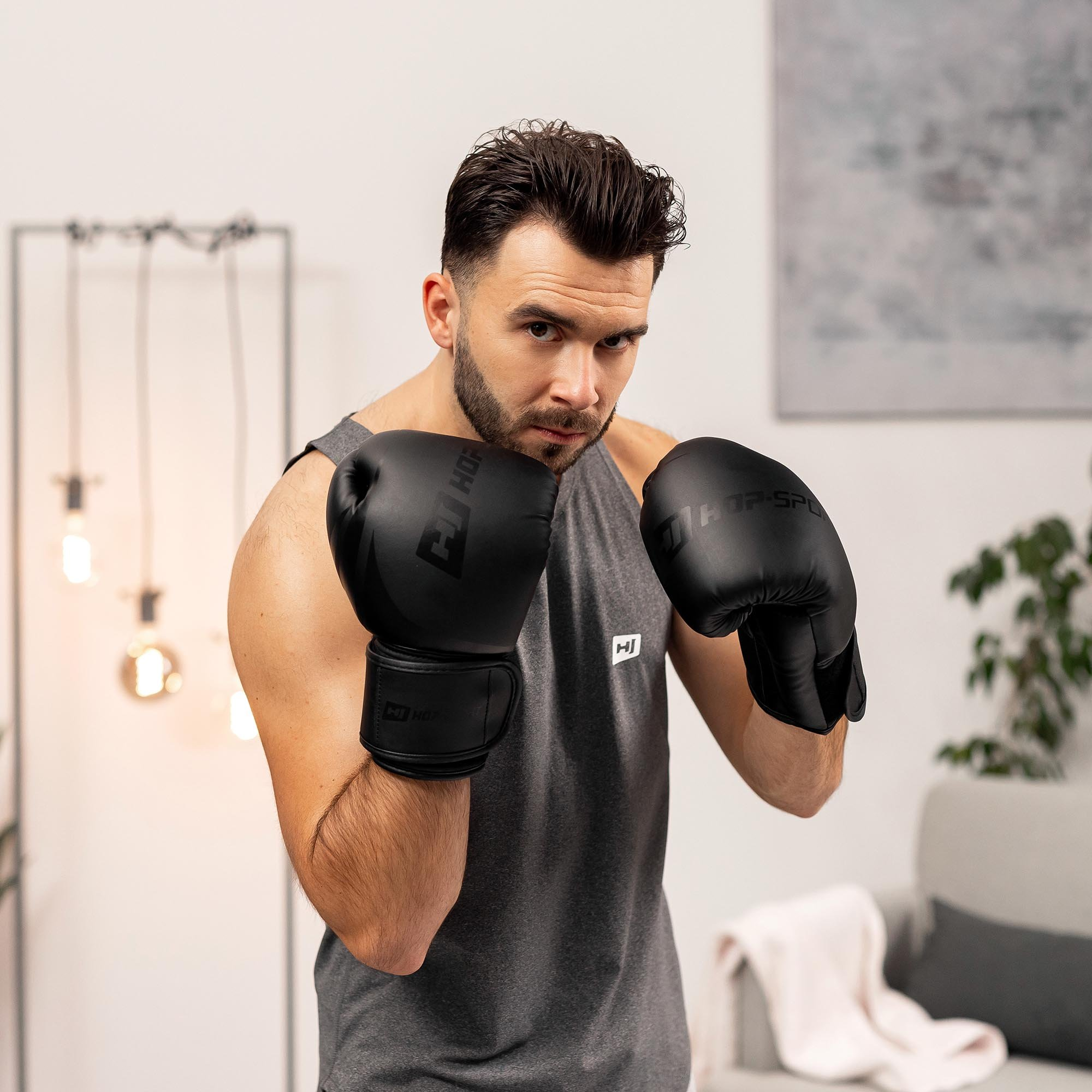 Ein Mann im grauen Tanktop und schwarzen Boxhandschuhen posiert in einem hellen Raum mit Lampe und Sofa im Hintergrund.