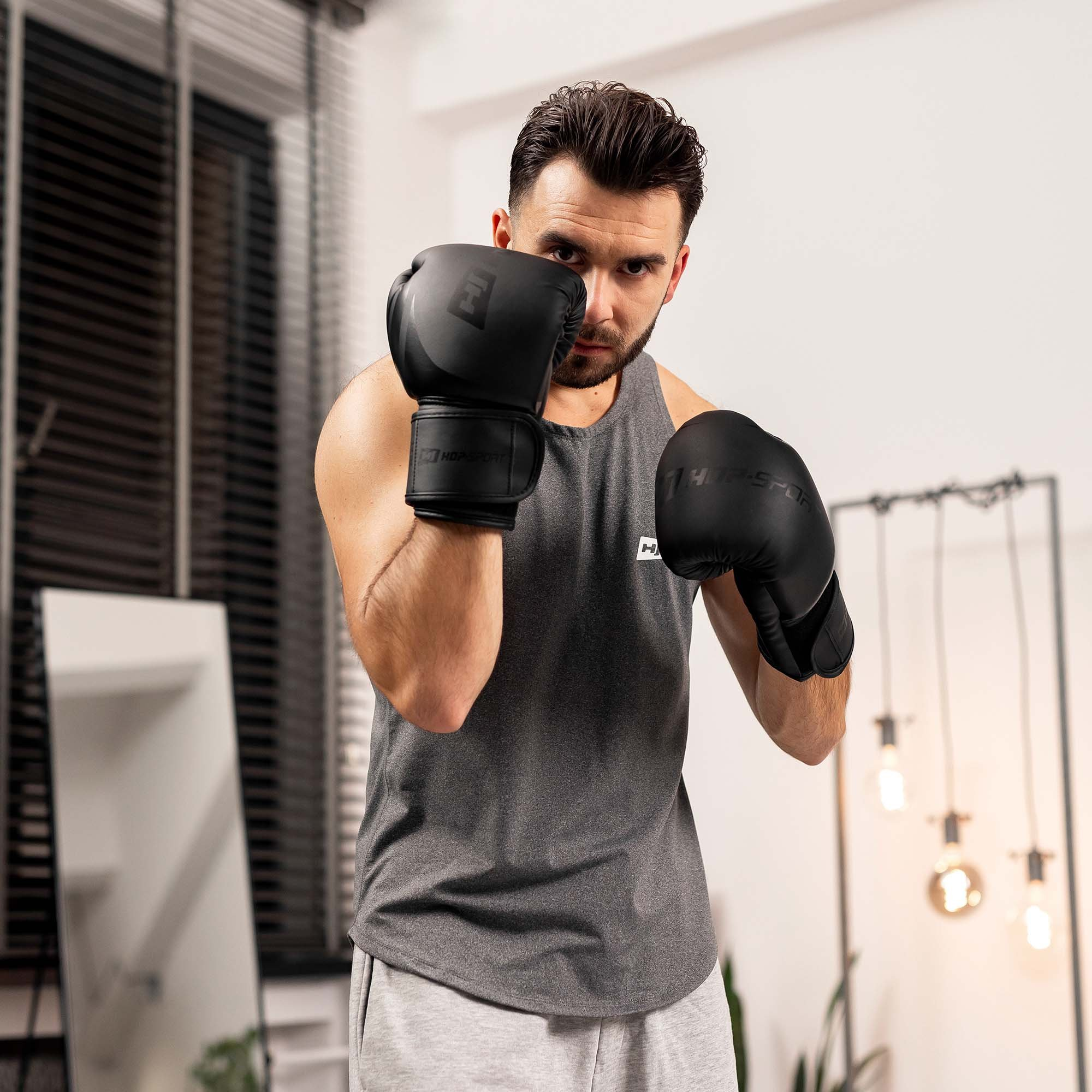 Ein Mann im grauen Tanktop und schwarzen Boxhandschuhen steht in einer Kampfhaltung in einem Raum.