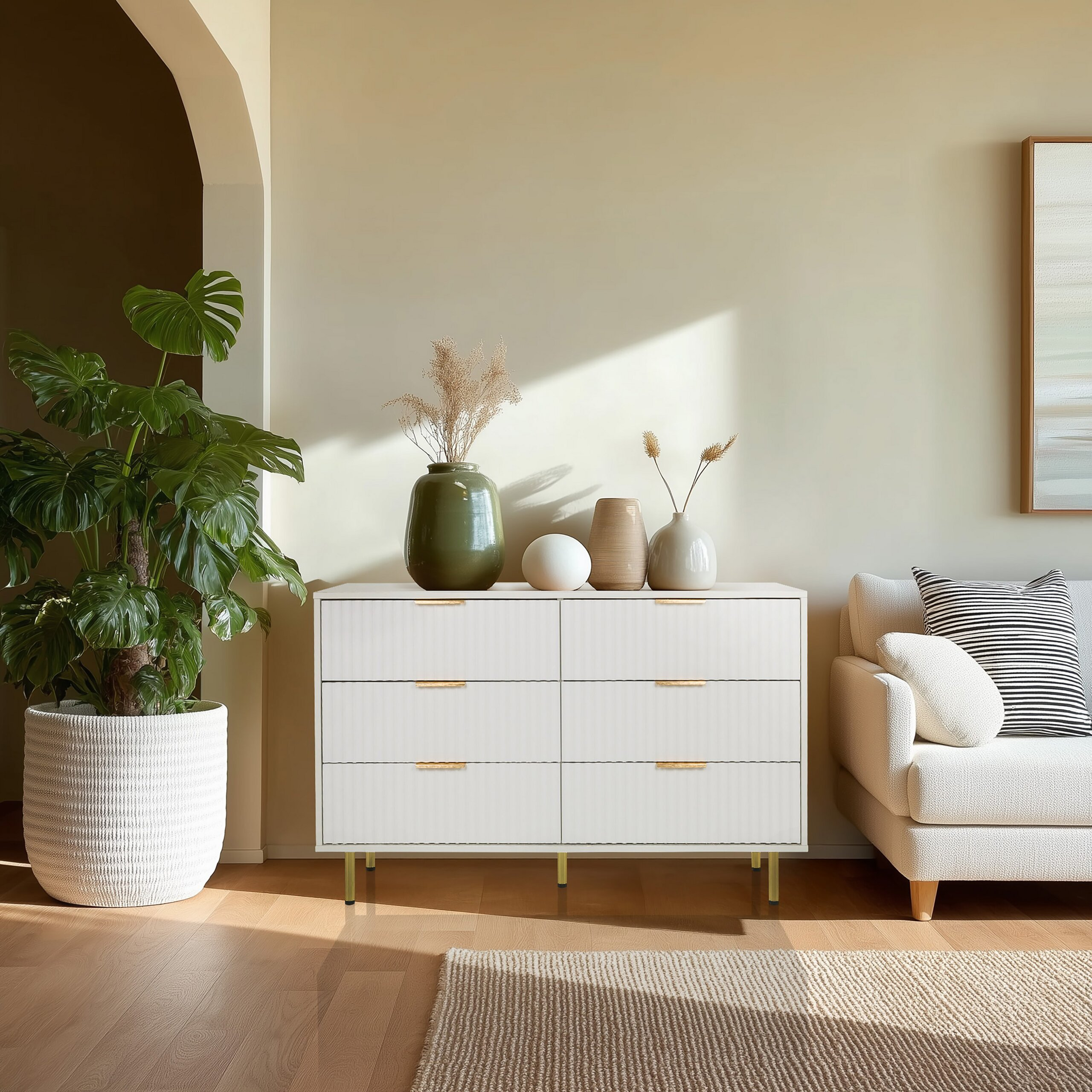Eine helle Kommode mit Vasen steht an einer beigen Wand, daneben eine große Topfpflanze und ein Sofa.