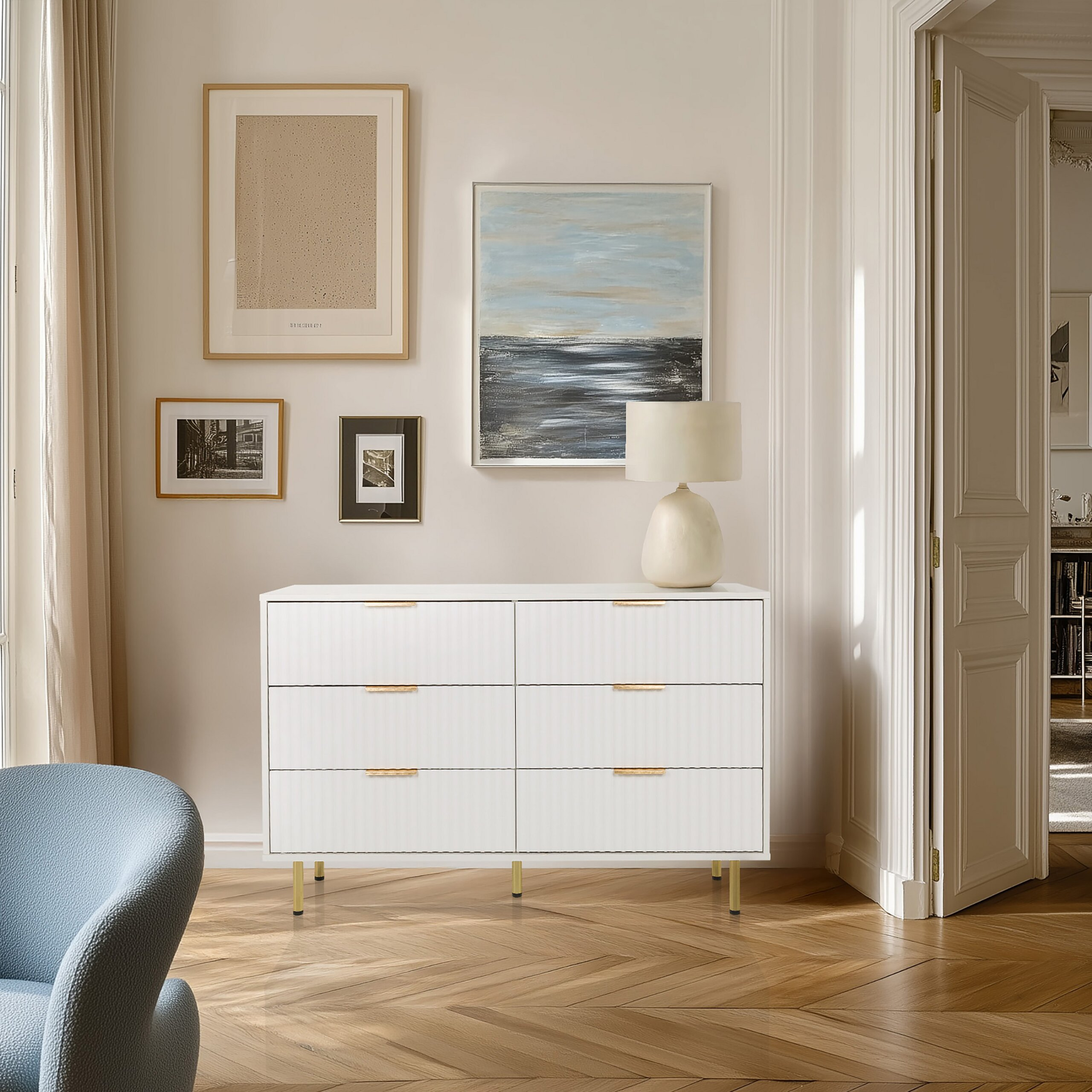 Eine weiße Kommode mit sechs Schubladen und goldenen Akzenten steht an einer Wand mit gerahmter Kunst und einer Lampe.