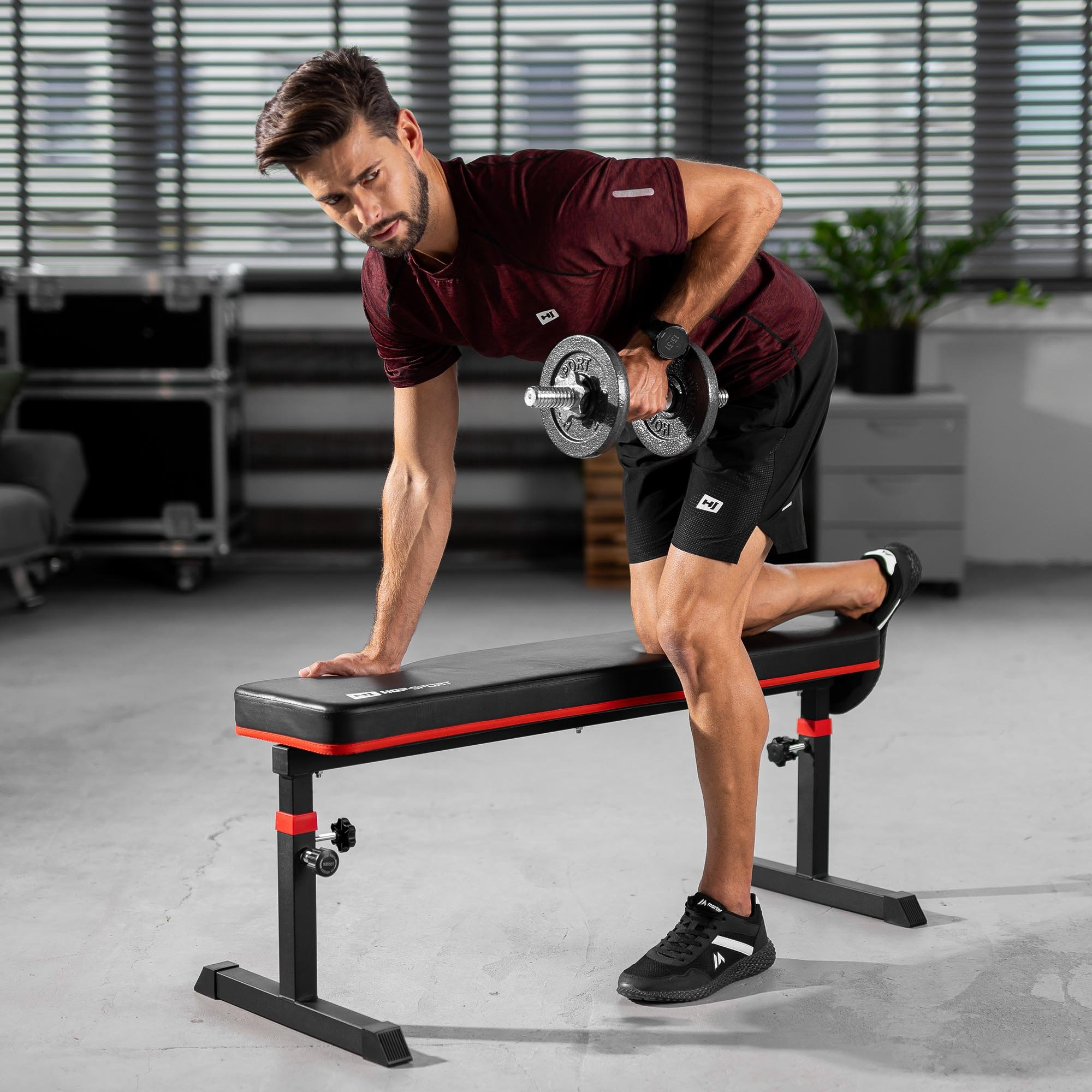 Ein Mann im roten T-Shirt und schwarzen Shorts trainiert mit einer Hantel auf einer Trainingsbank.