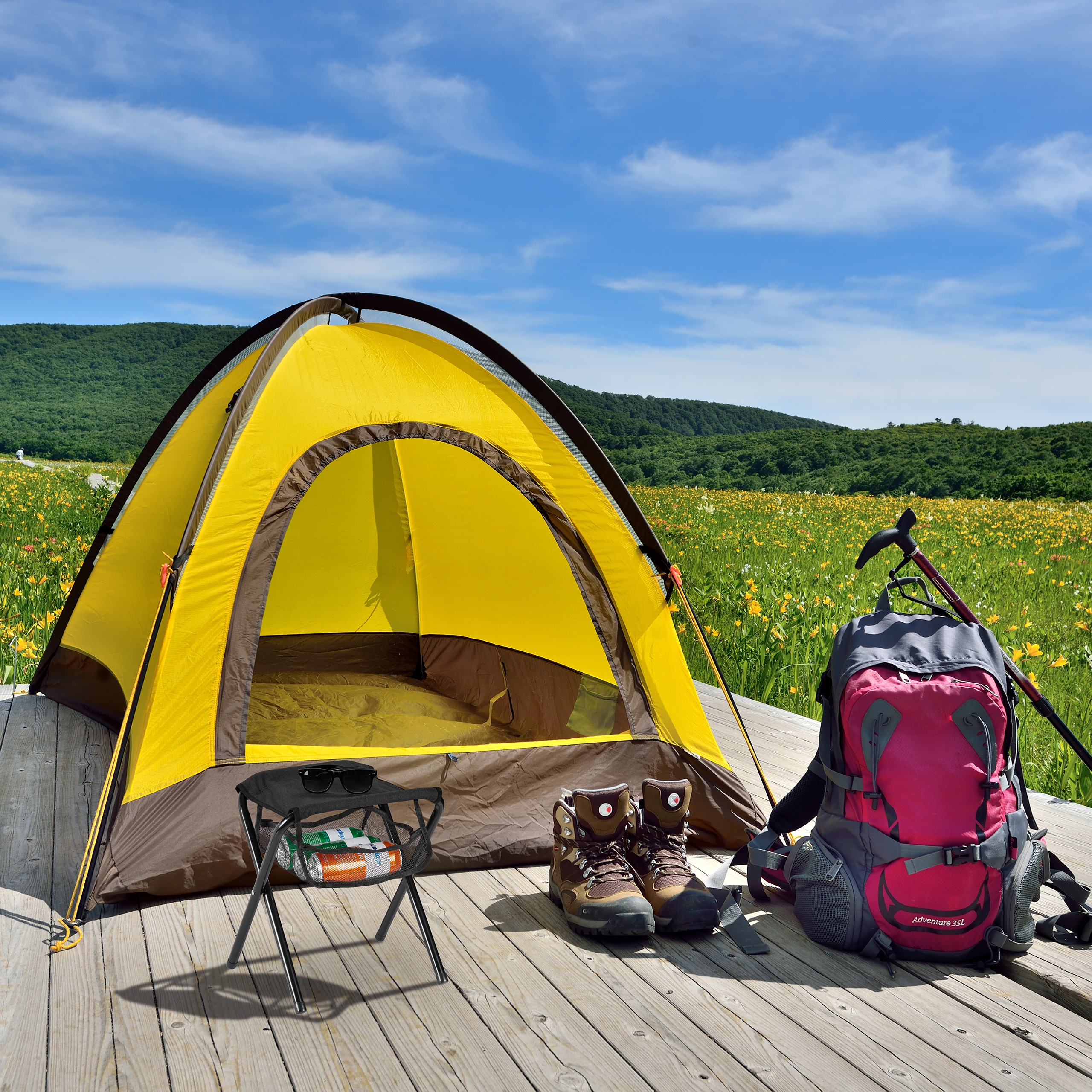 Gelbes Zelt, Rucksack, Wanderschuhe und ein Stuhl auf Holzplanken vor einer Wiese.