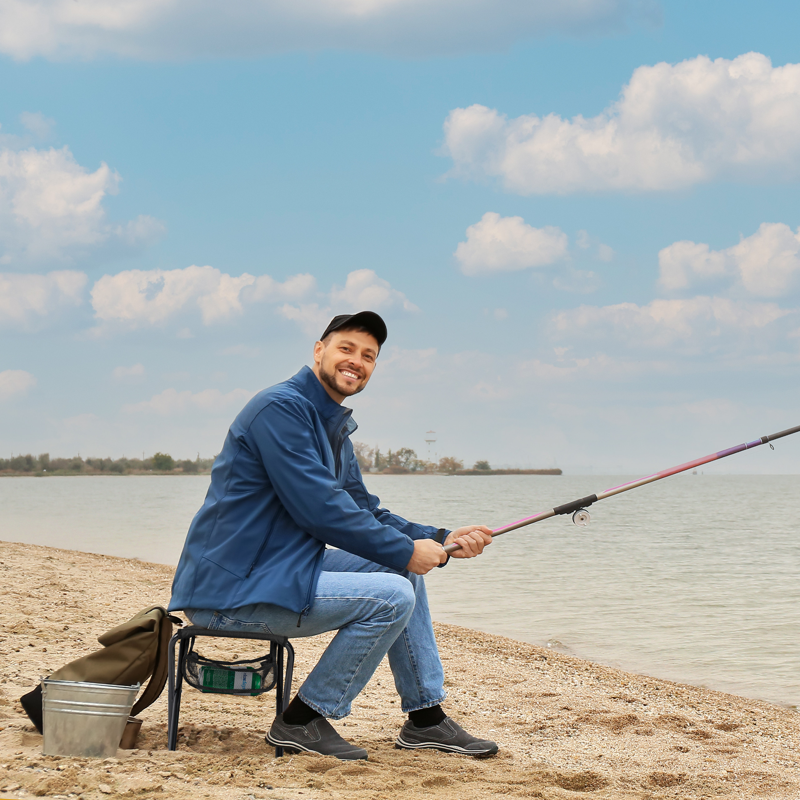 Ein lächelnder Mann sitzt auf einem Stuhl am See und hält eine Angelrute in der Hand.