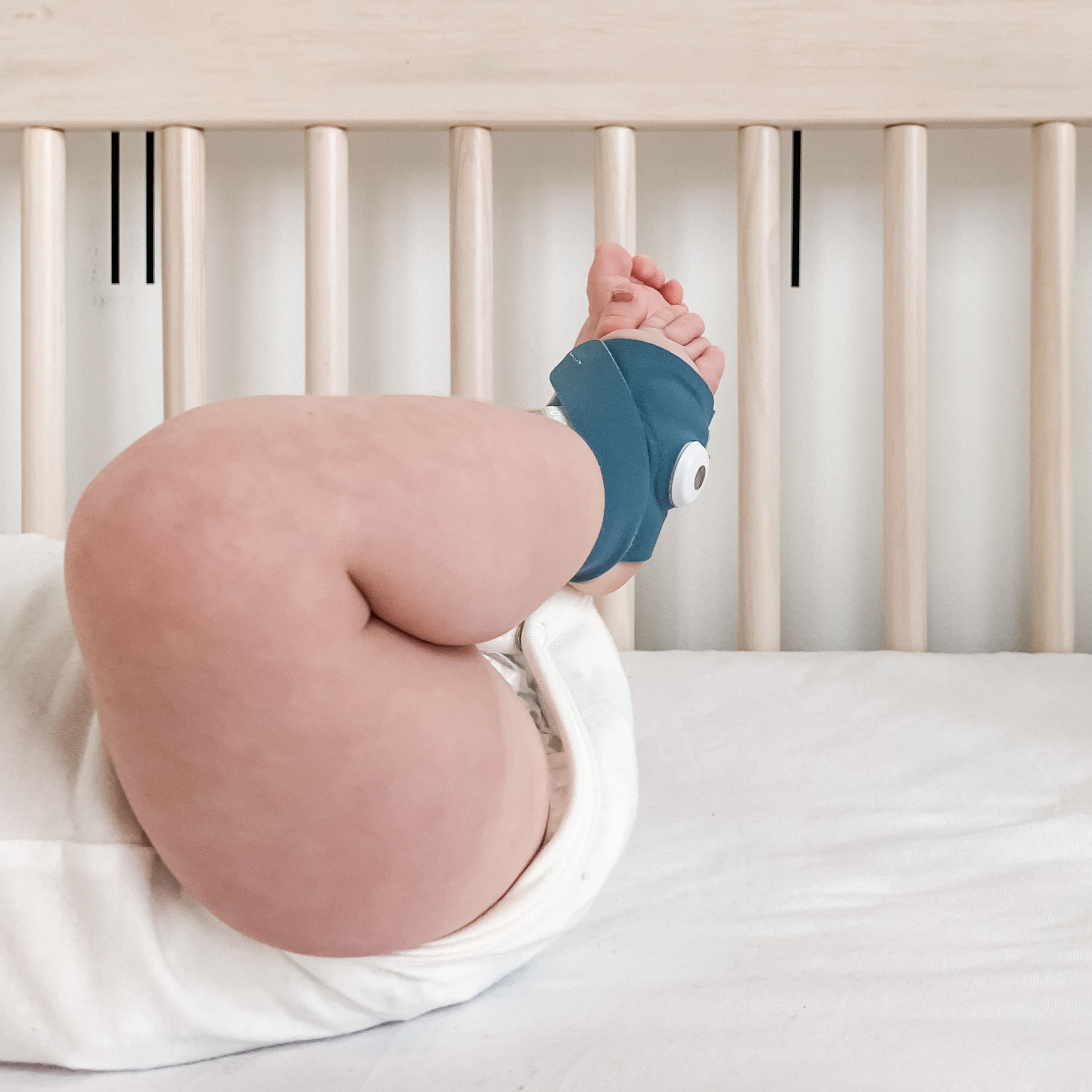 Der Fuß eines Babys in einer blauen Monitorsocke, das in einem Bettchen mit weißer Bettwäsche liegt.