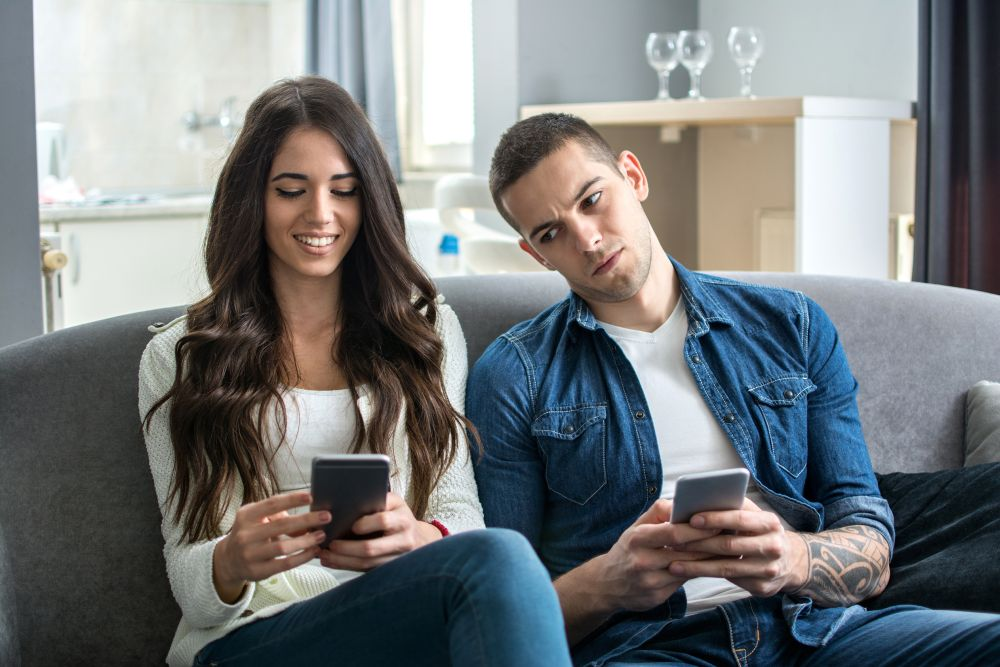 Kobieta uśmiecha się do telefonu, podczas gdy siedzący obok niej mężczyzna z ciekawością zagląda.