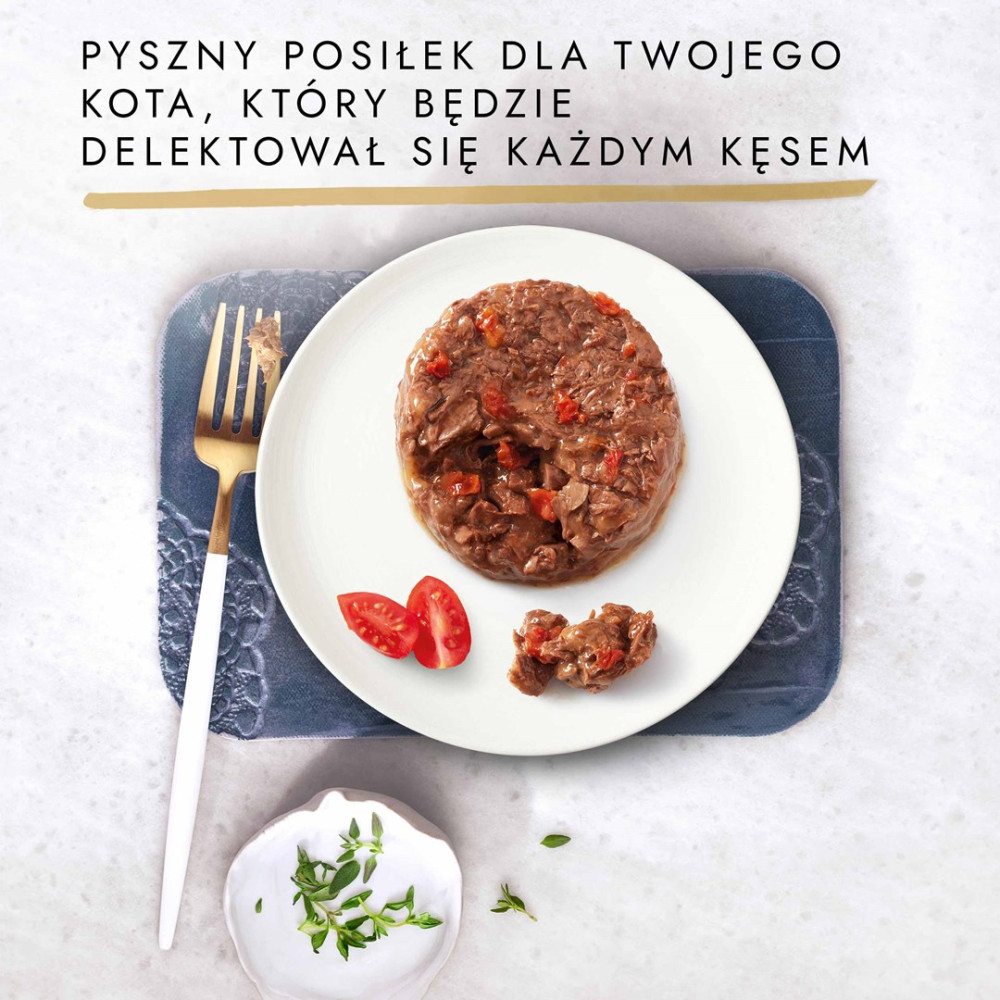 Kocia karma na białym talerzu z kawałkami pomidora, złotym widelcem i ziołami na szarym tle.