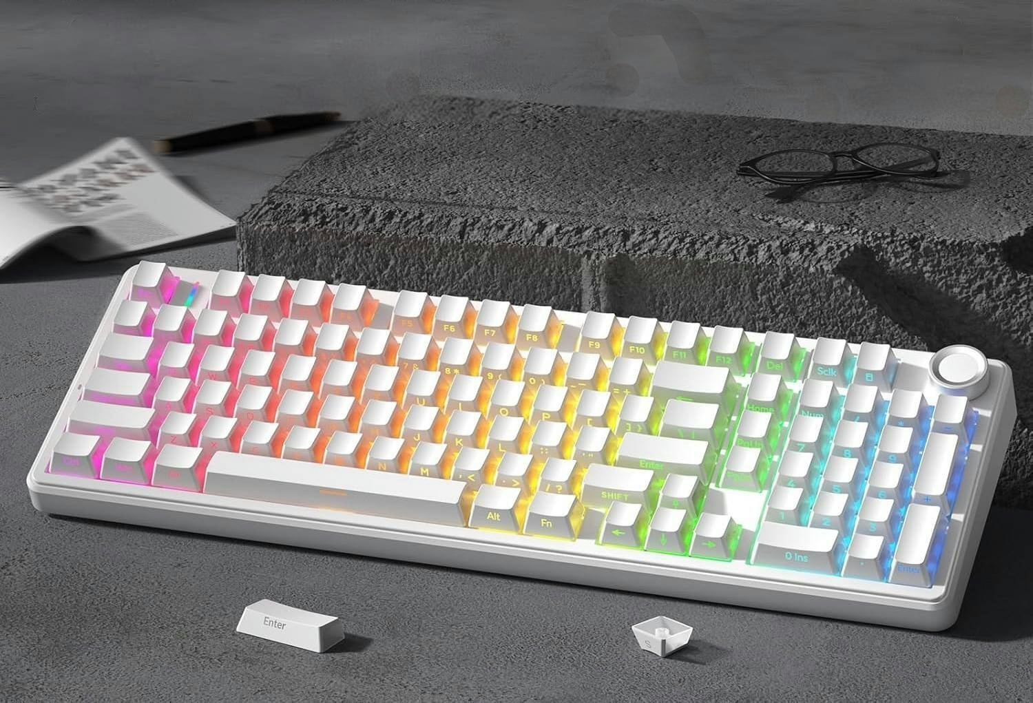 Biała klawiatura mechaniczna z podświetleniem RGB na kamiennym stole, z okularami i notatkami w tle.