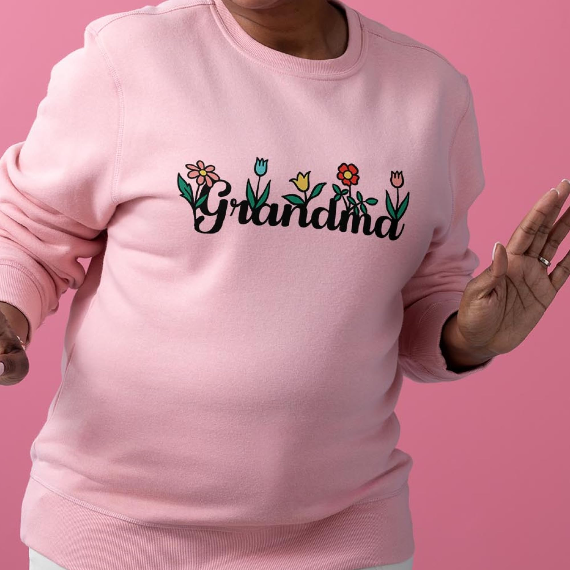Une personne porte un sweat rose avec le mot "Grandma" en écriture noire entrelacé de fleurs colorées.