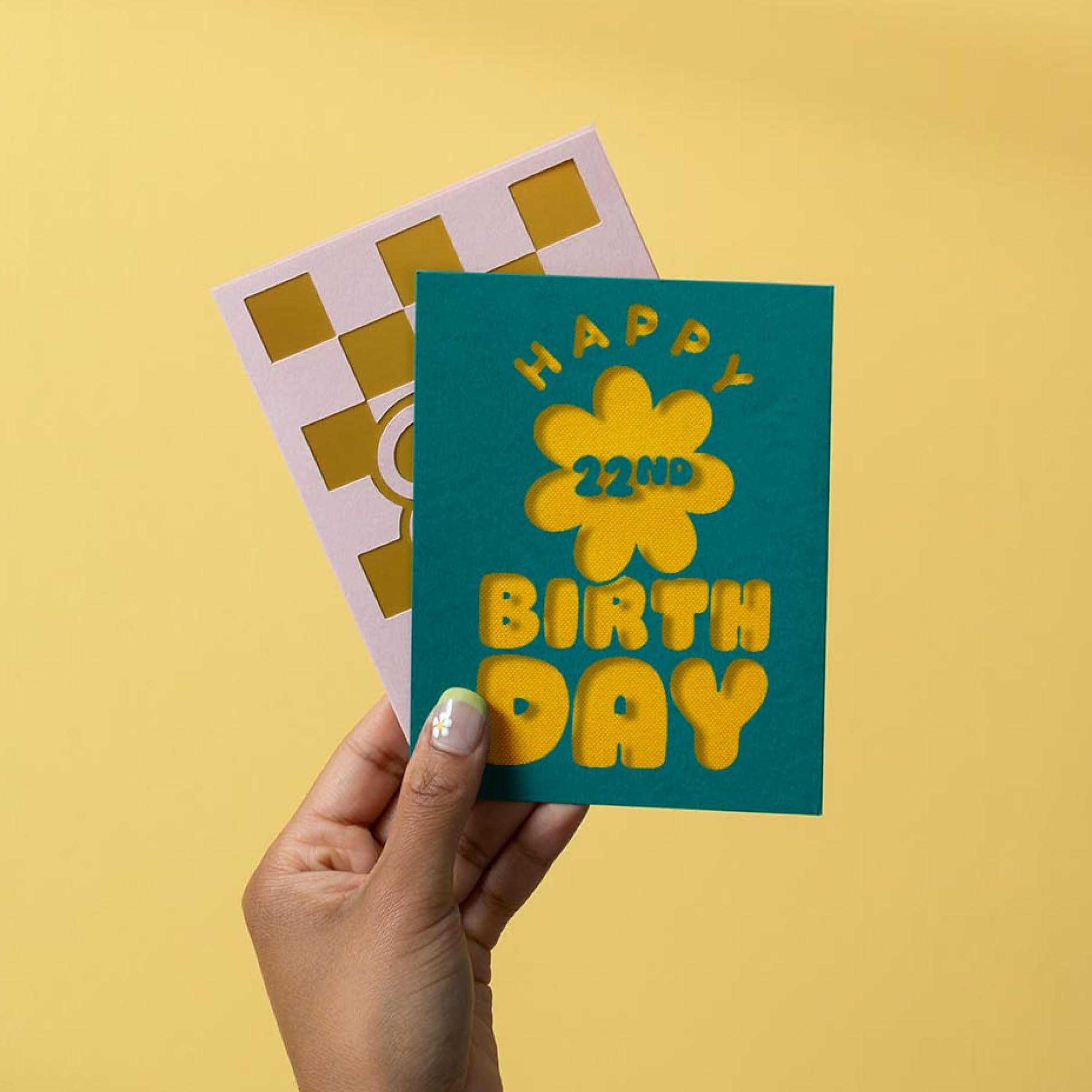 Une main tient deux cartes d'anniversaire sur fond jaune, celle de devant est verte avec une inscription jaune 22nd Birthday.