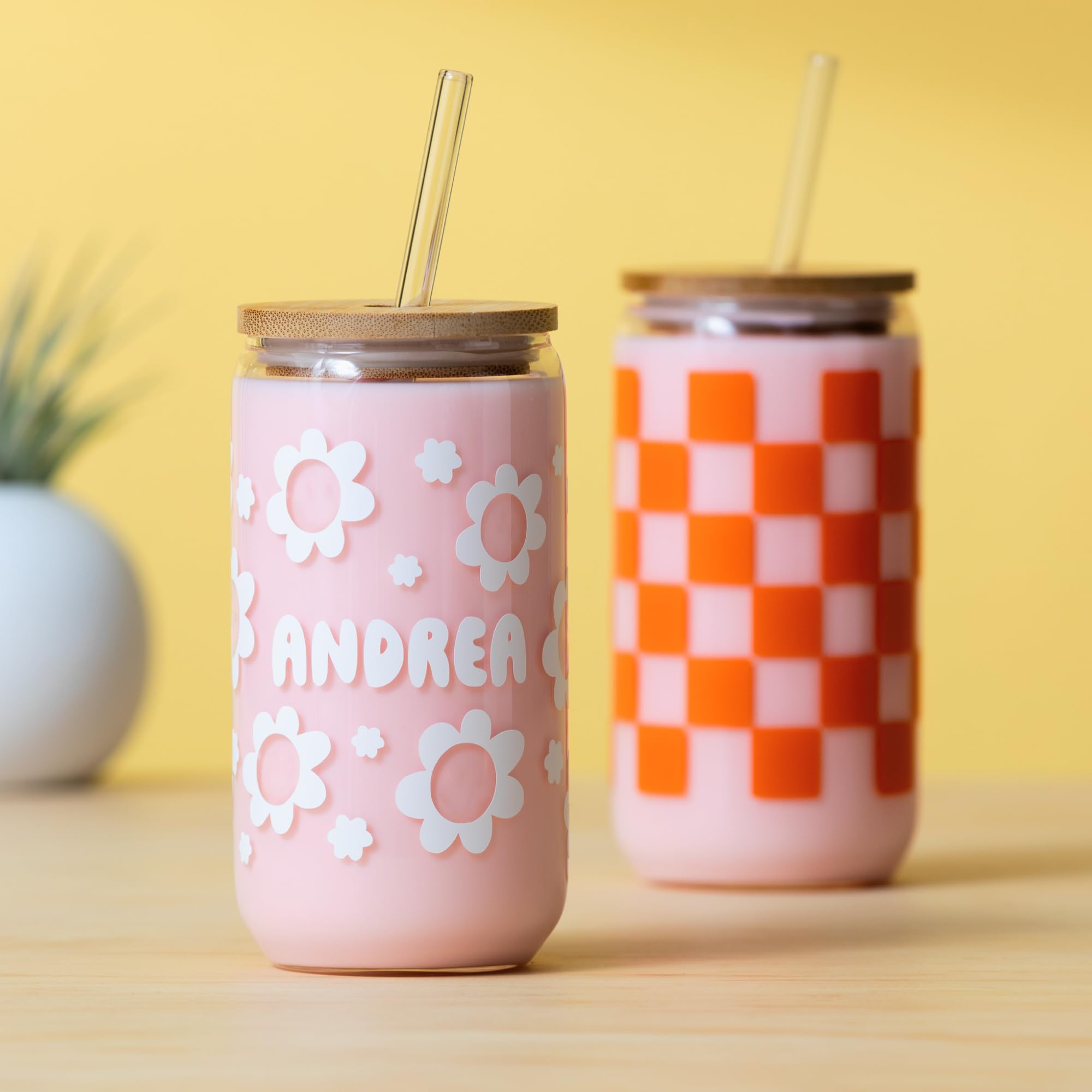 Deux tasses en verre rose avec des couvercles en bambou et des pailles sont sur une table en bois.