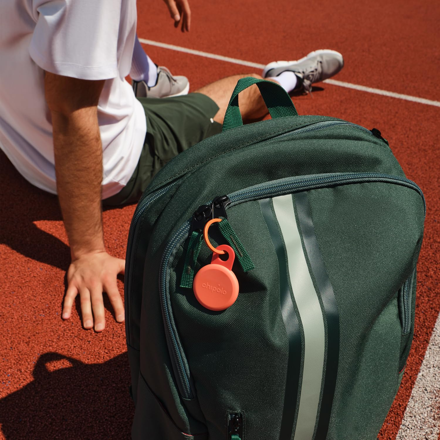 Ein grüner Rucksack mit orangefarbenem Schlüsselfinder liegt auf einer roten Laufbahn neben einem sitzenden Athleten.