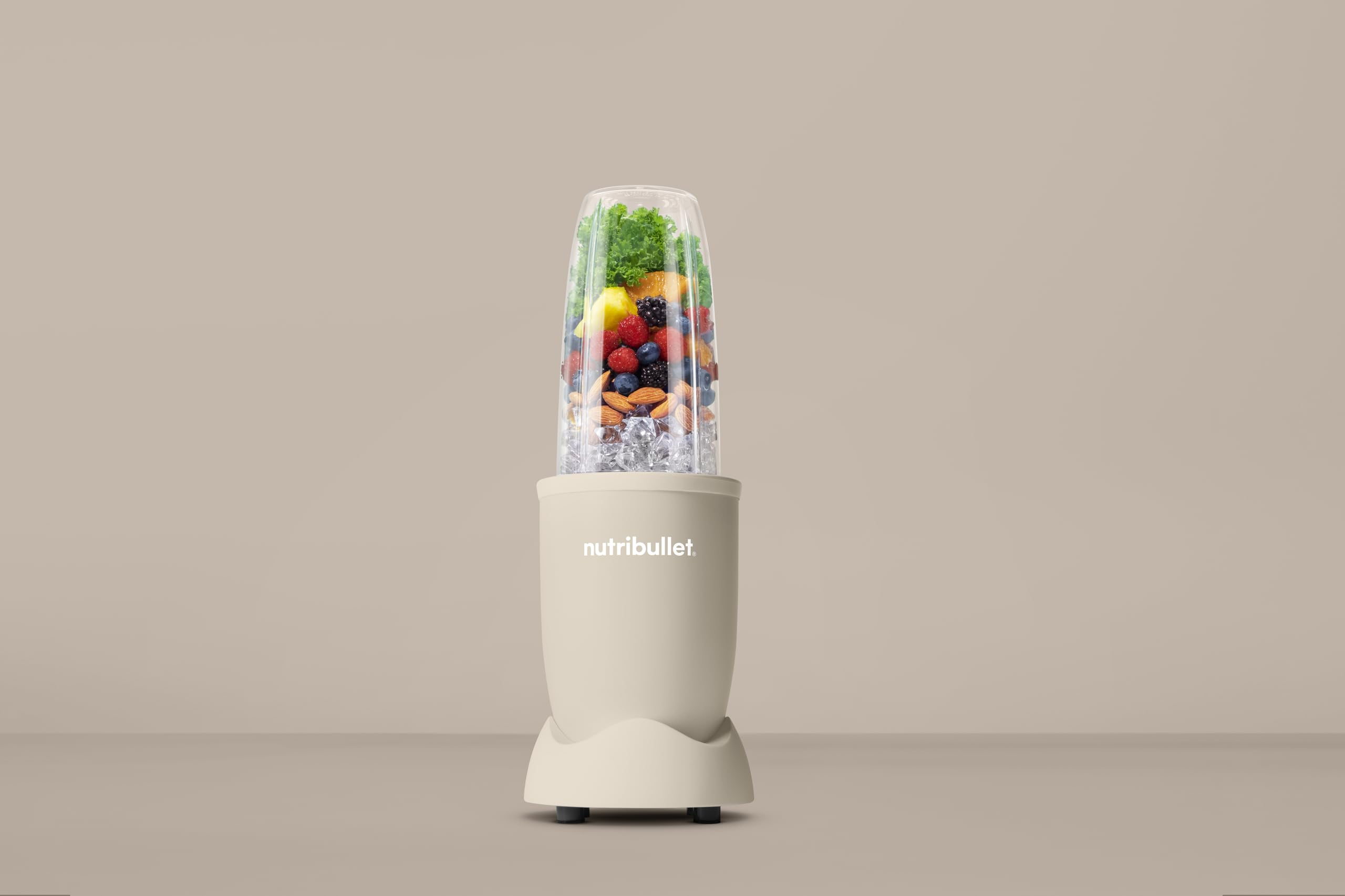Ein beiger Nutribullet-Mixer gefüllt mit Eis, Nüssen, Beeren und grünem Blattgemüse vor beigem Hintergrund.
