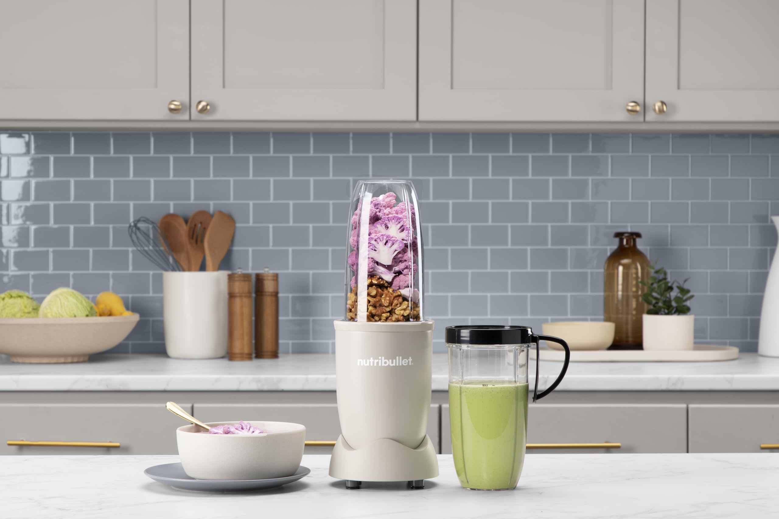 Ein Beigeblender mit Walnüssen und violettem Blumenkohl steht auf der Küchenarbeitsplatte neben einem grünen Smoothie.