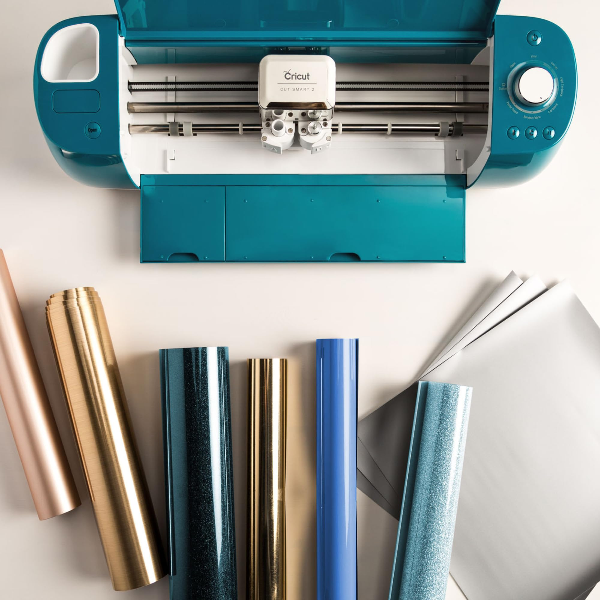 Machine de découpe Cricut sarcelle avec divers rouleaux de vinyle et de papier colorés disposés devant.