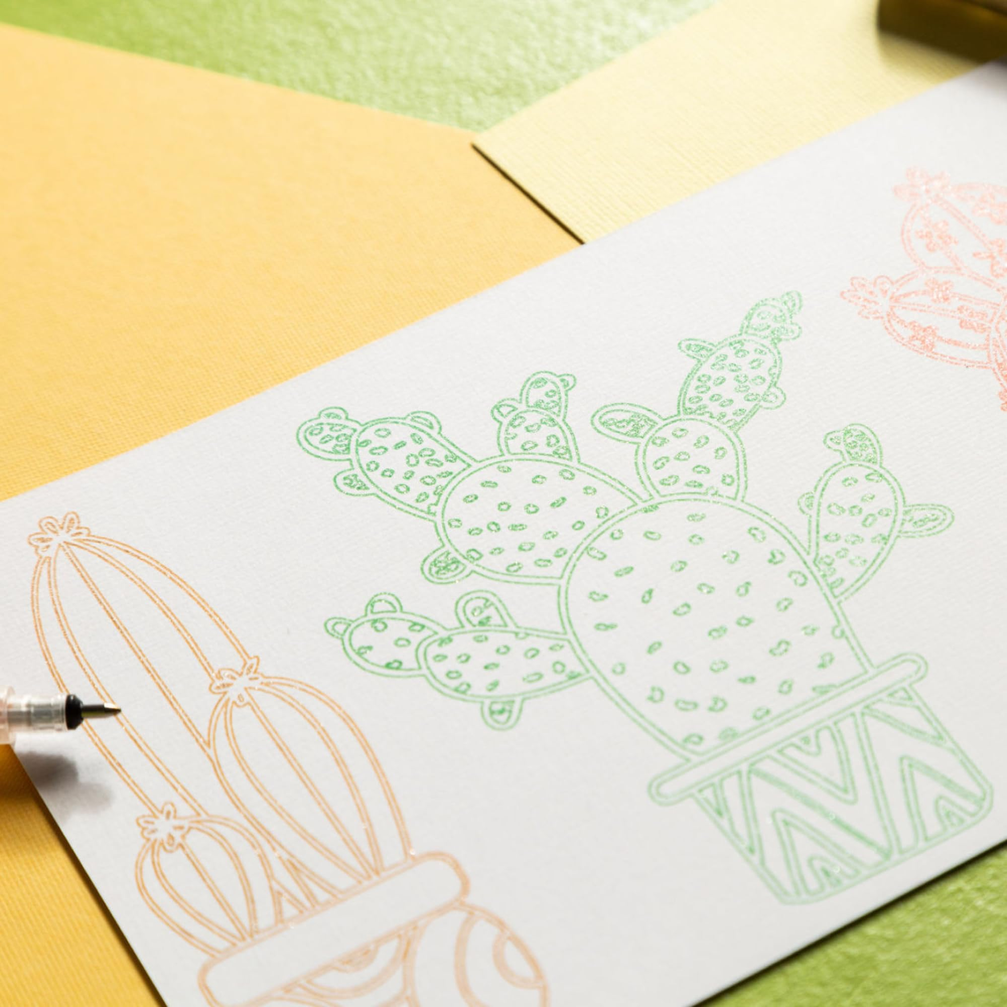 Un papier blanc avec des dessins de cactus verts, orange et roses repose sur un fond coloré avec une pointe de stylo visible.