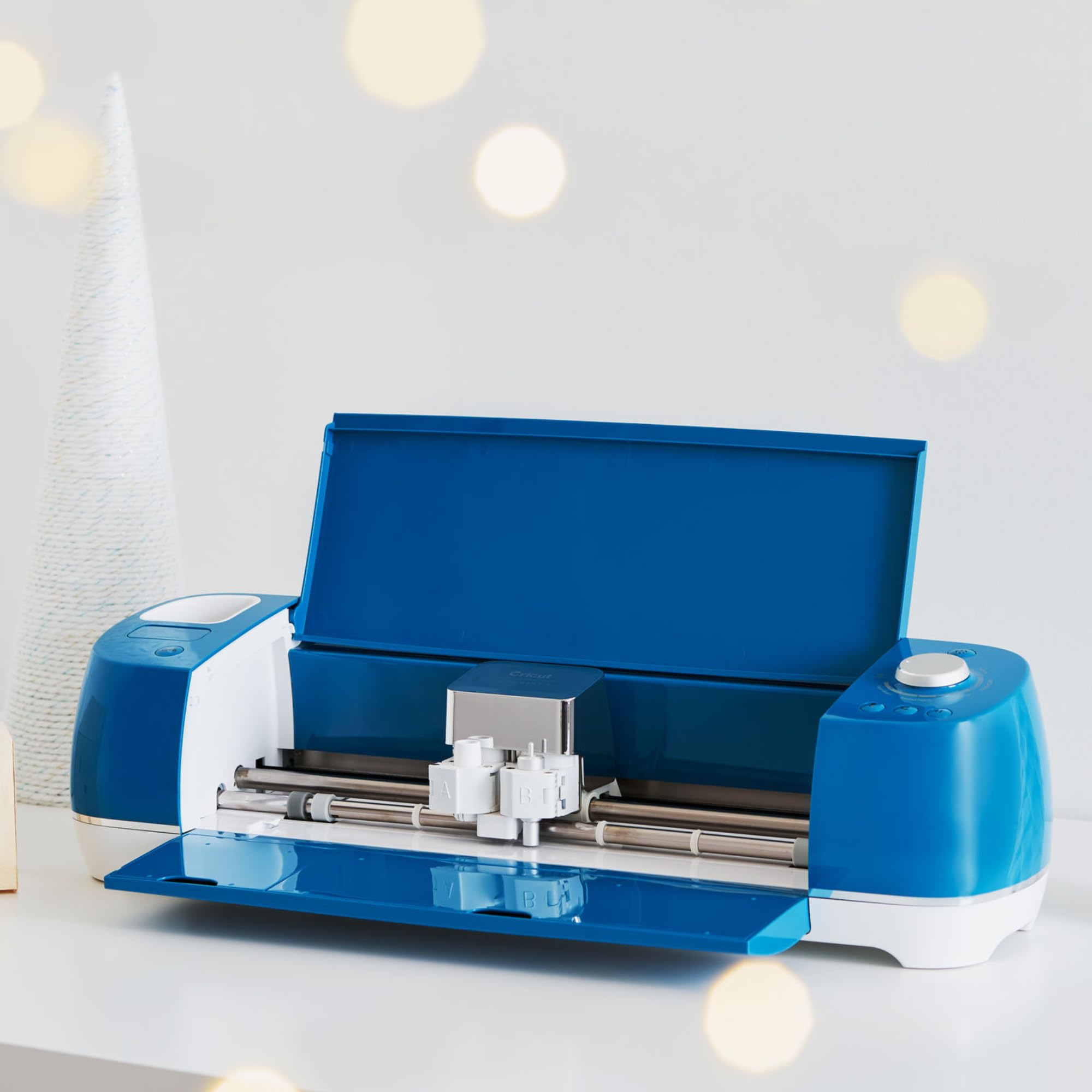 Une machine de découpe Cricut bleue et blanche repose sur une surface blanche, avec un petit arbre décoratif blanc en arrière-plan.