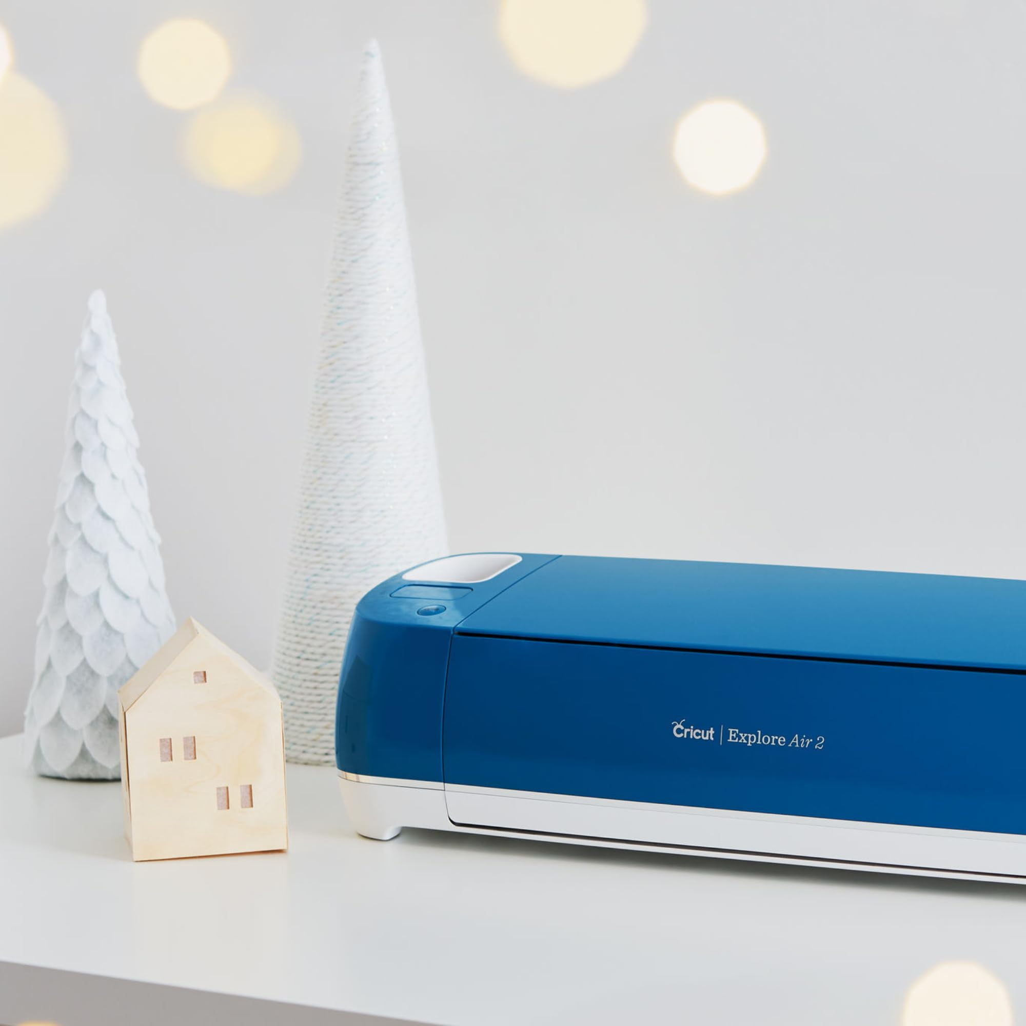 Une imprimante bleue Cricut Explore Air 2 à côté de deux sapins de Noël blancs et d'une maison en bois.