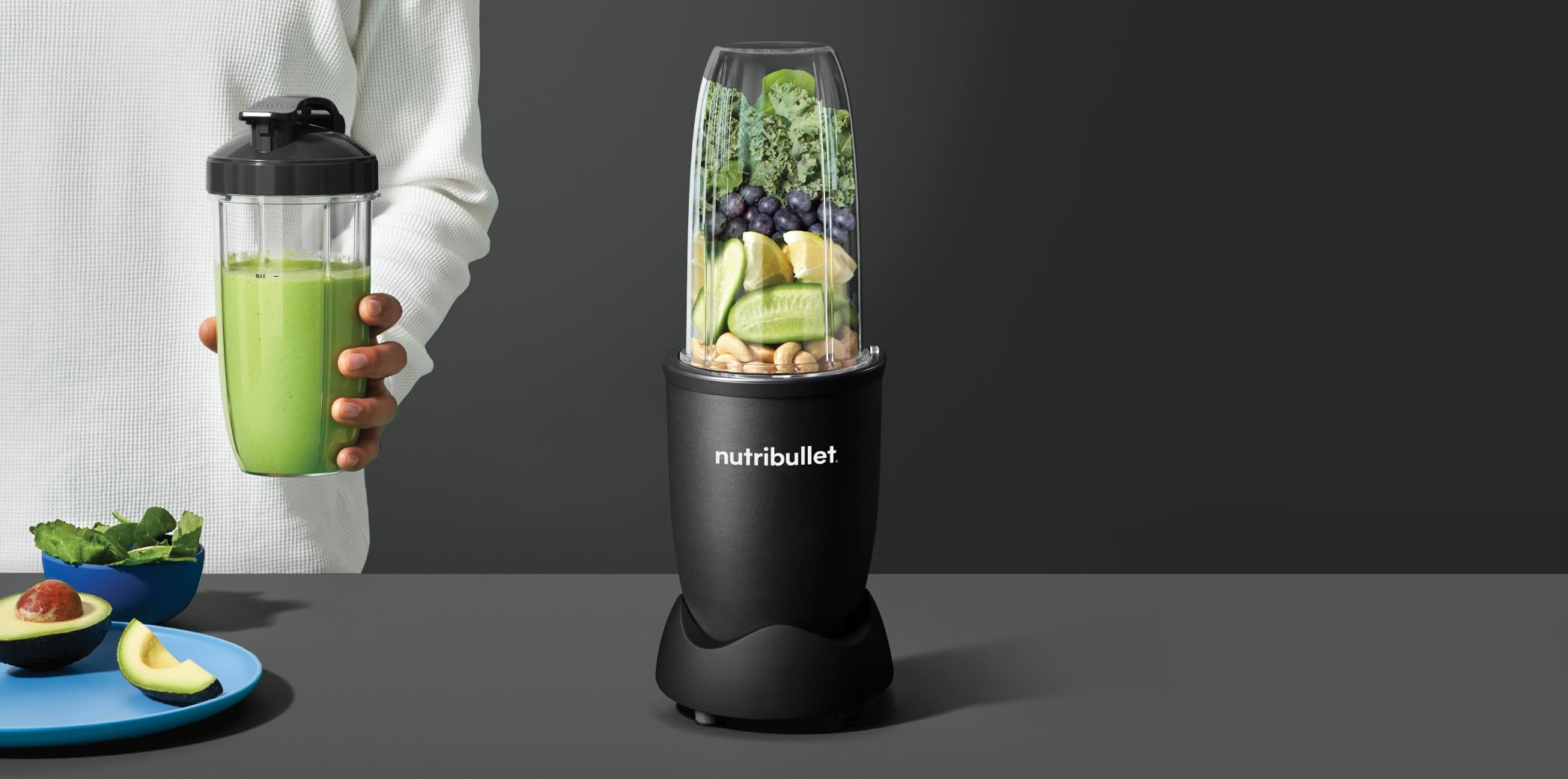 Ein schwarzer Nutribullet-Mixer mit Gemüse und Beeren, neben einer Person, die einen grünen Smoothie hält.