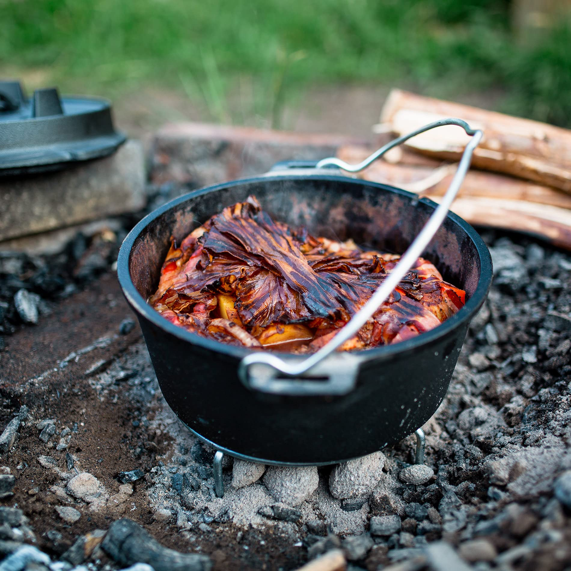 Une cocotte en fonte noire sur des braises, remplie de nourriture enveloppée de bacon croustillant.
