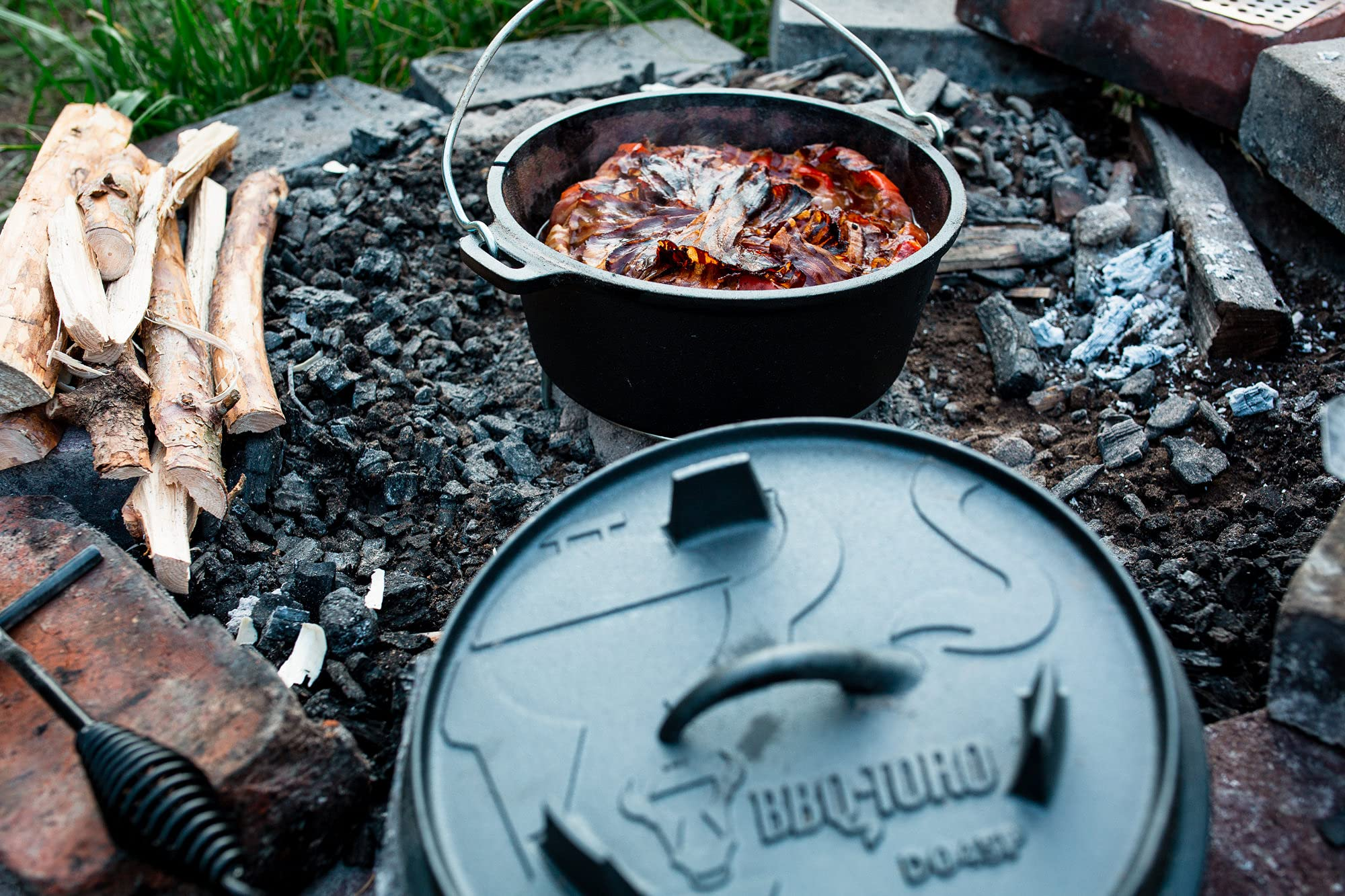 Une cocotte en fonte noire remplie de nourriture repose sur des braises près de bois de chauffage.
