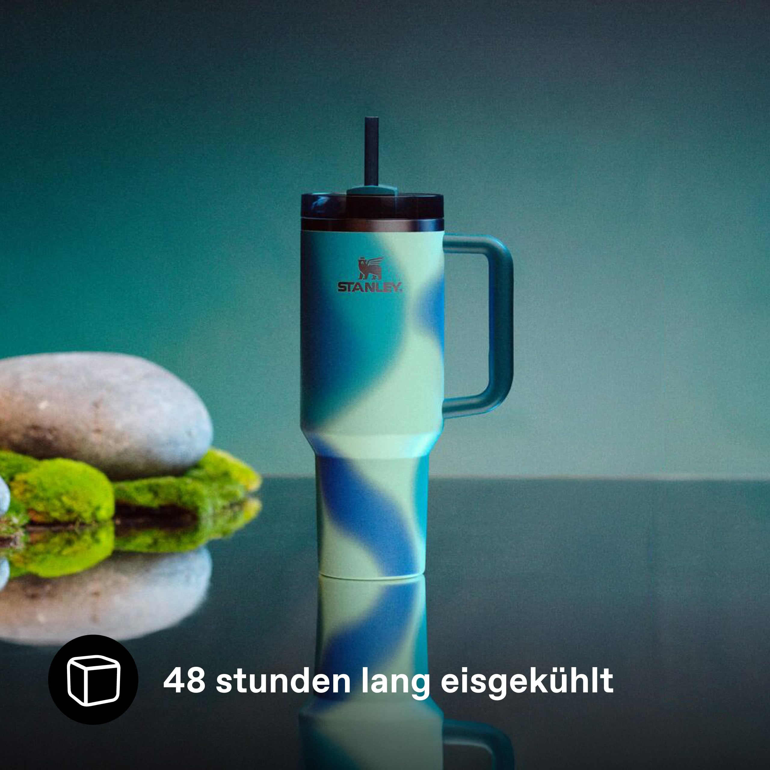 Ein blau-grün gemusterter Stanley-Trinkbecher mit Strohhalm steht auf einem glänzenden Tisch neben Moos und Stein.