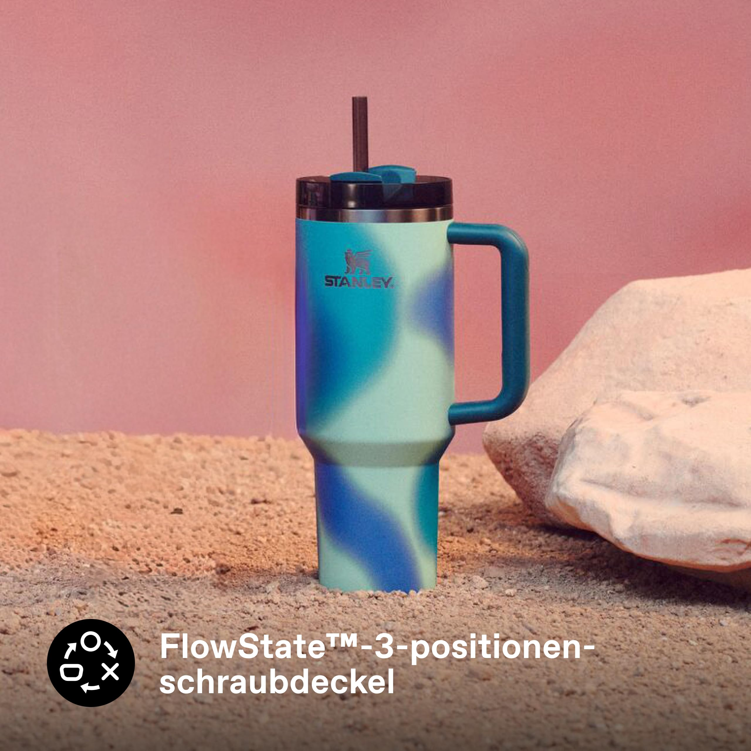 Ein blau-grüner Stanley-Becher mit Strohhalm steht auf sandigem Boden neben einem Felsen vor rosa Hintergrund.