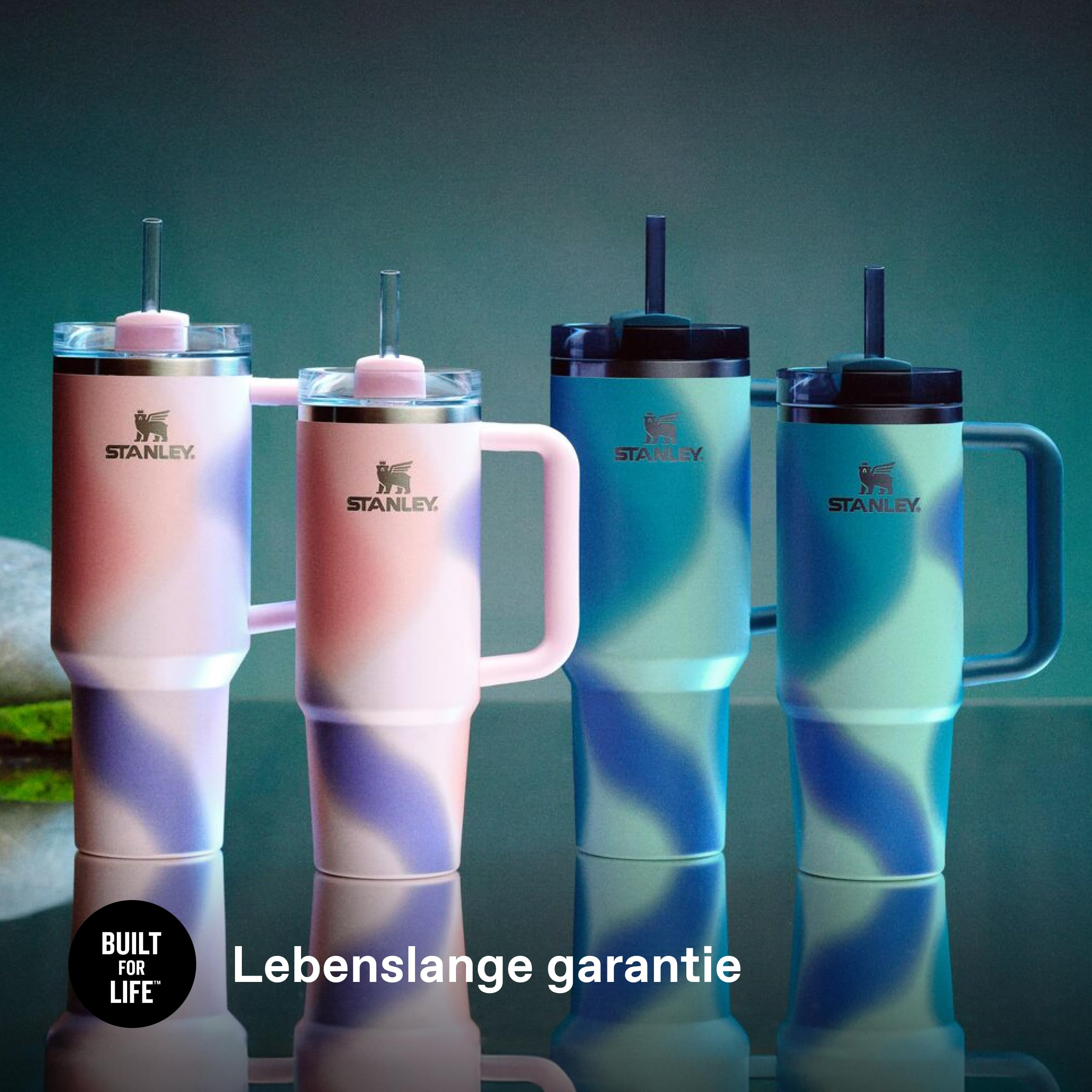 Vier Stanley-Becher mit Strohhalmen in Rosa und Blau stehen auf einer reflektierenden Oberfläche vor dunklem Hintergrund.