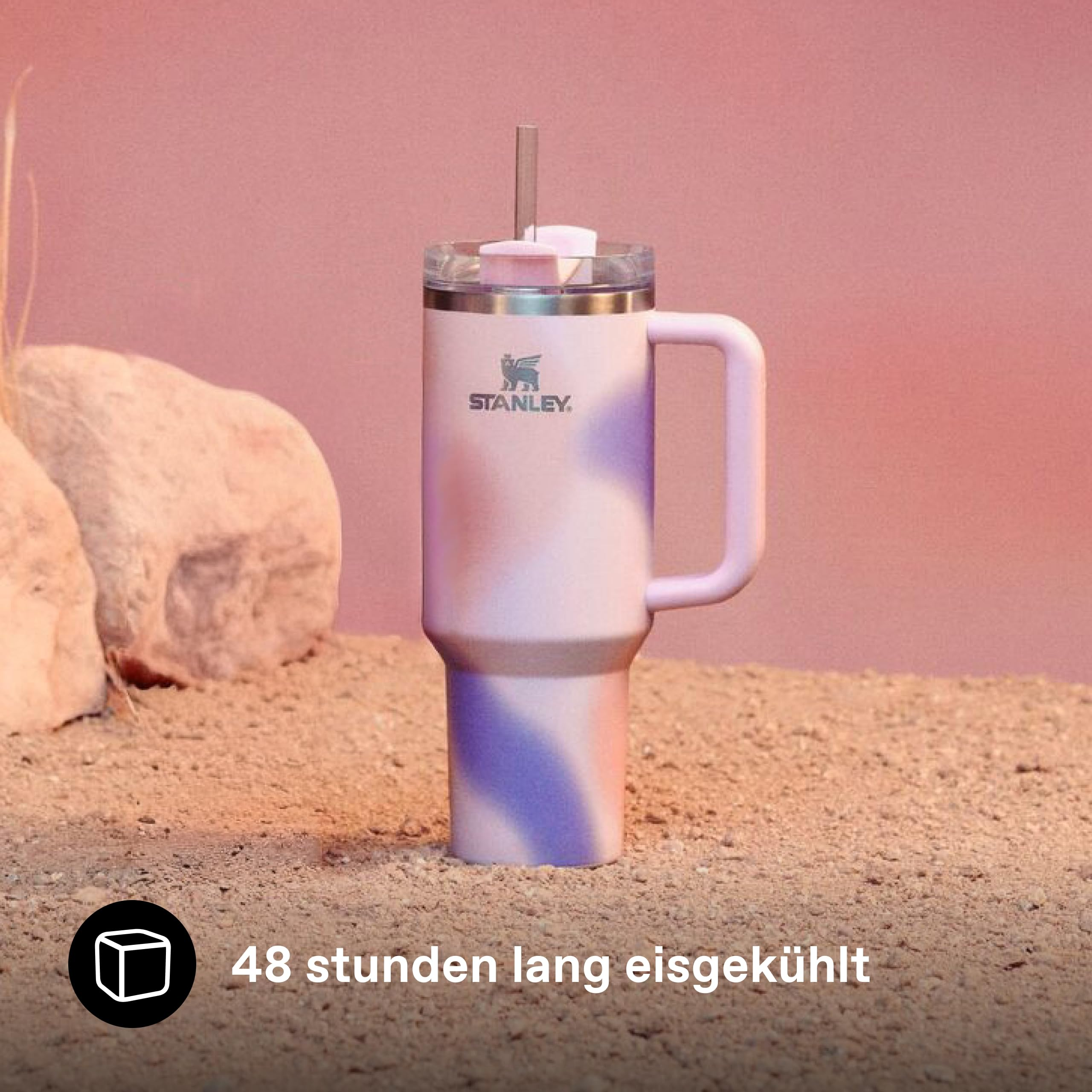 Ein pastellvioletter und -rosafarbener Stanley-Becher mit Strohhalm steht auf Sand vor rosa Hintergrund, Steine dahinter.