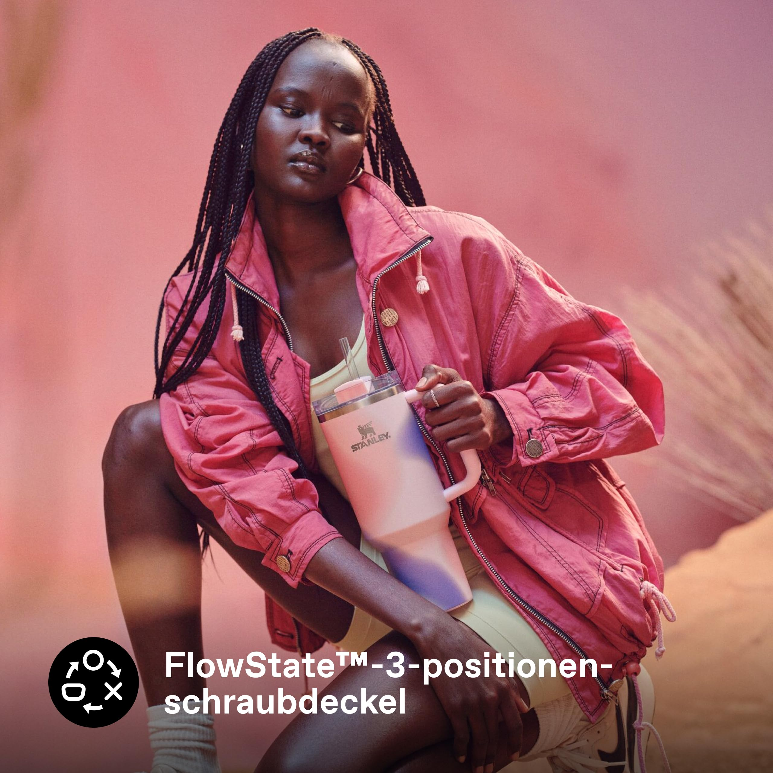 Eine Frau in pinker Jacke hält einen Stanley-Trinkbecher in einer warmen, staubigen Landschaft.