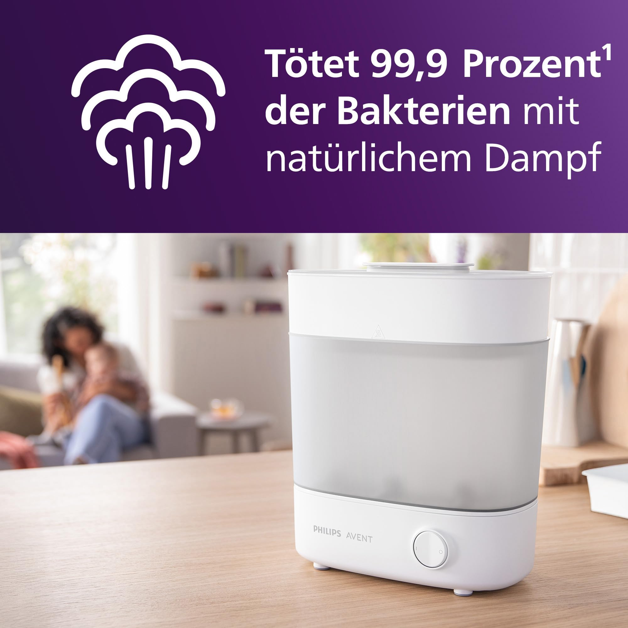 Ein weißer Philips Avent Dampfsterilisator steht auf einer Holztheke; im Hintergrund sitzt eine Mutter mit Kind.