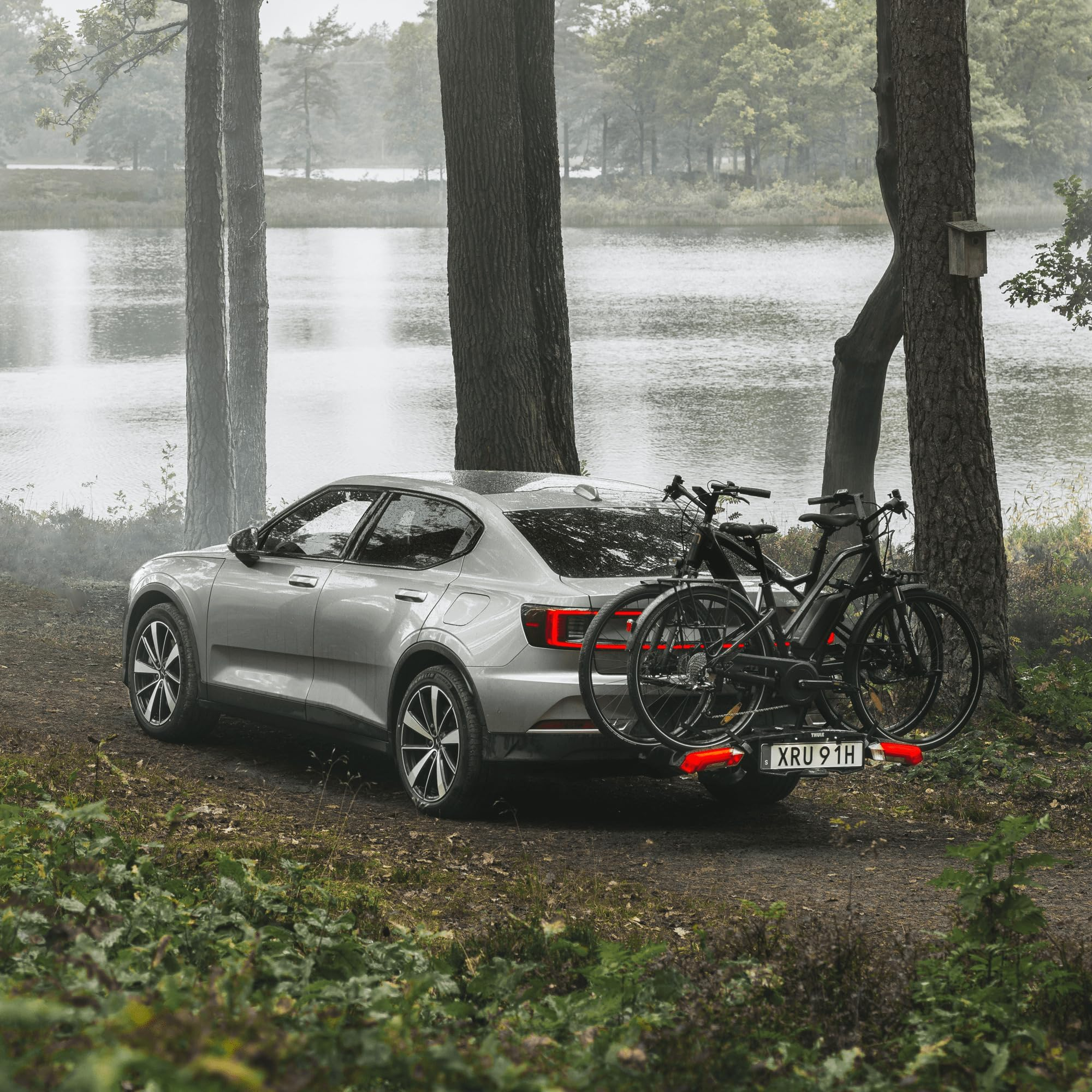 Un'auto grigia con portabici e due biciclette parcheggiata su un sentiero vicino a un lago.