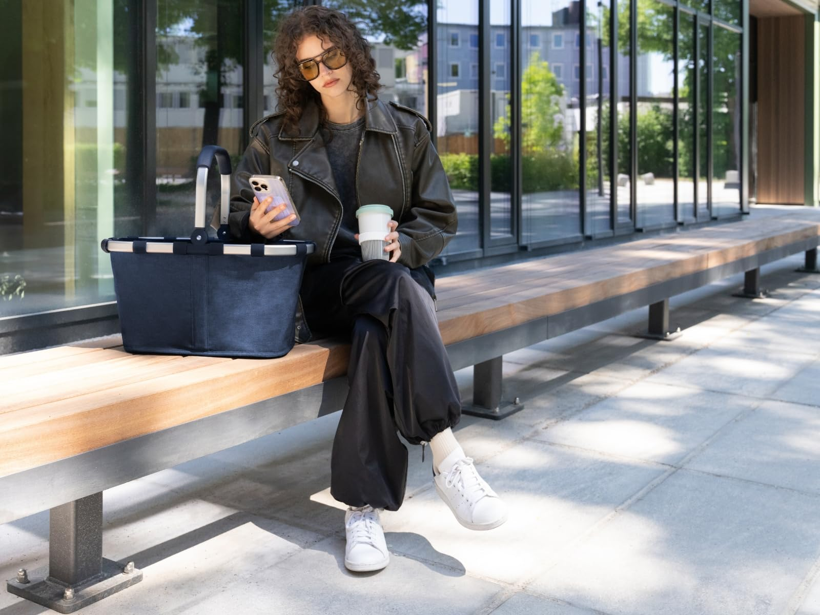 Eine Frau in schwarzer Lederjacke sitzt mit Handy und Kaffee auf einer Bank neben einer blauen Tasche vor einem Gebäude.