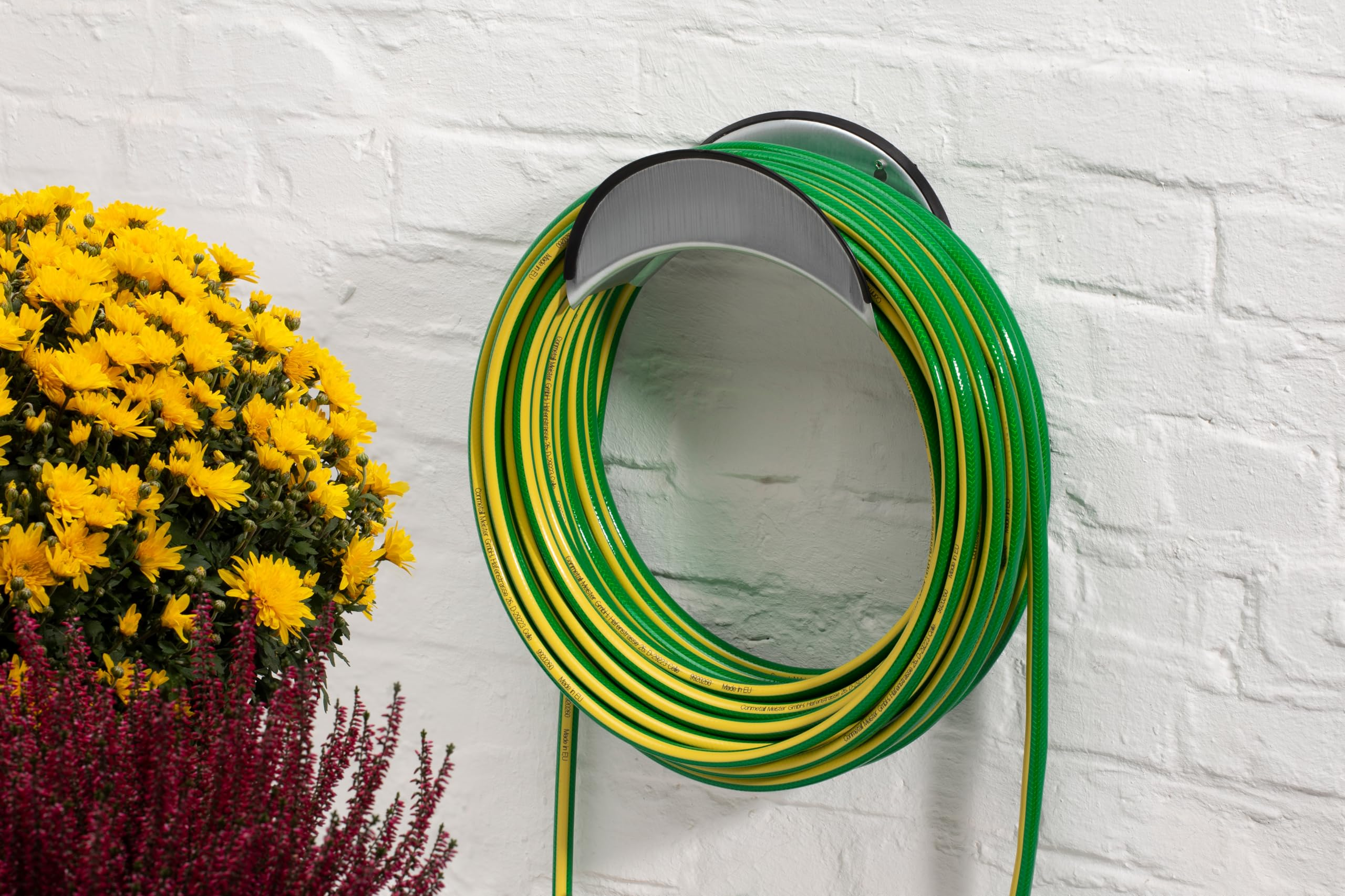 Ein grün-gelber Gartenschlauch auf einem Metallhalter, neben gelben und lila Blumen an einer weißen Backsteinwand.