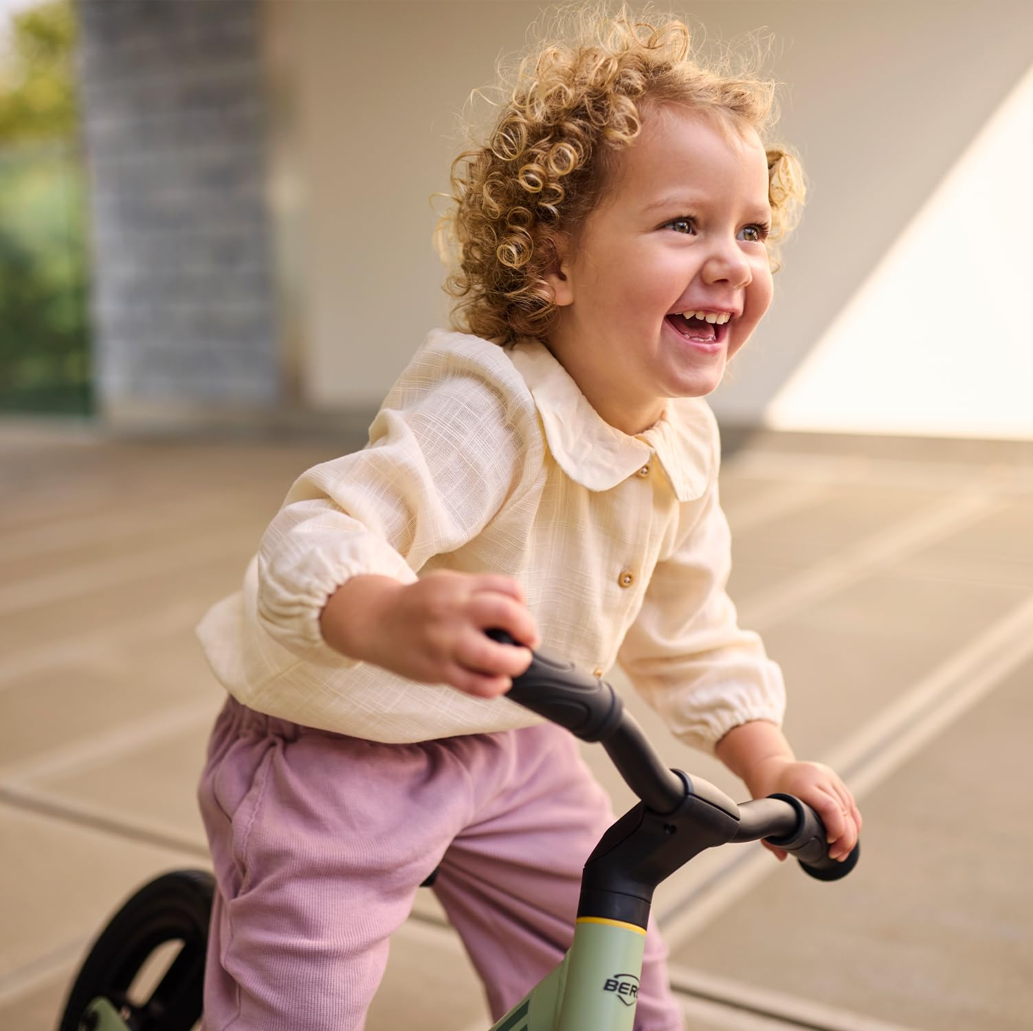 Un bambino sorridente con i capelli biondi ricci va in bici senza pedali all'aperto, indossando una camicetta crema.