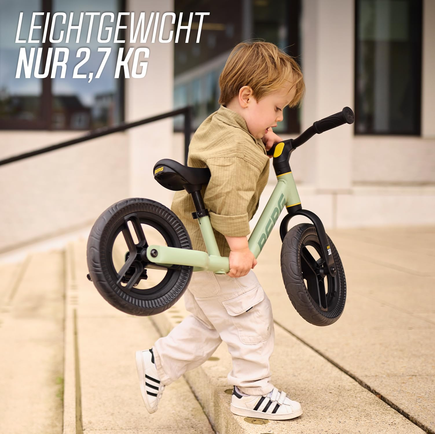 Bambino con vestiti cachi trasporta una bici senza pedali verde chiaro giù per le scale esterne.