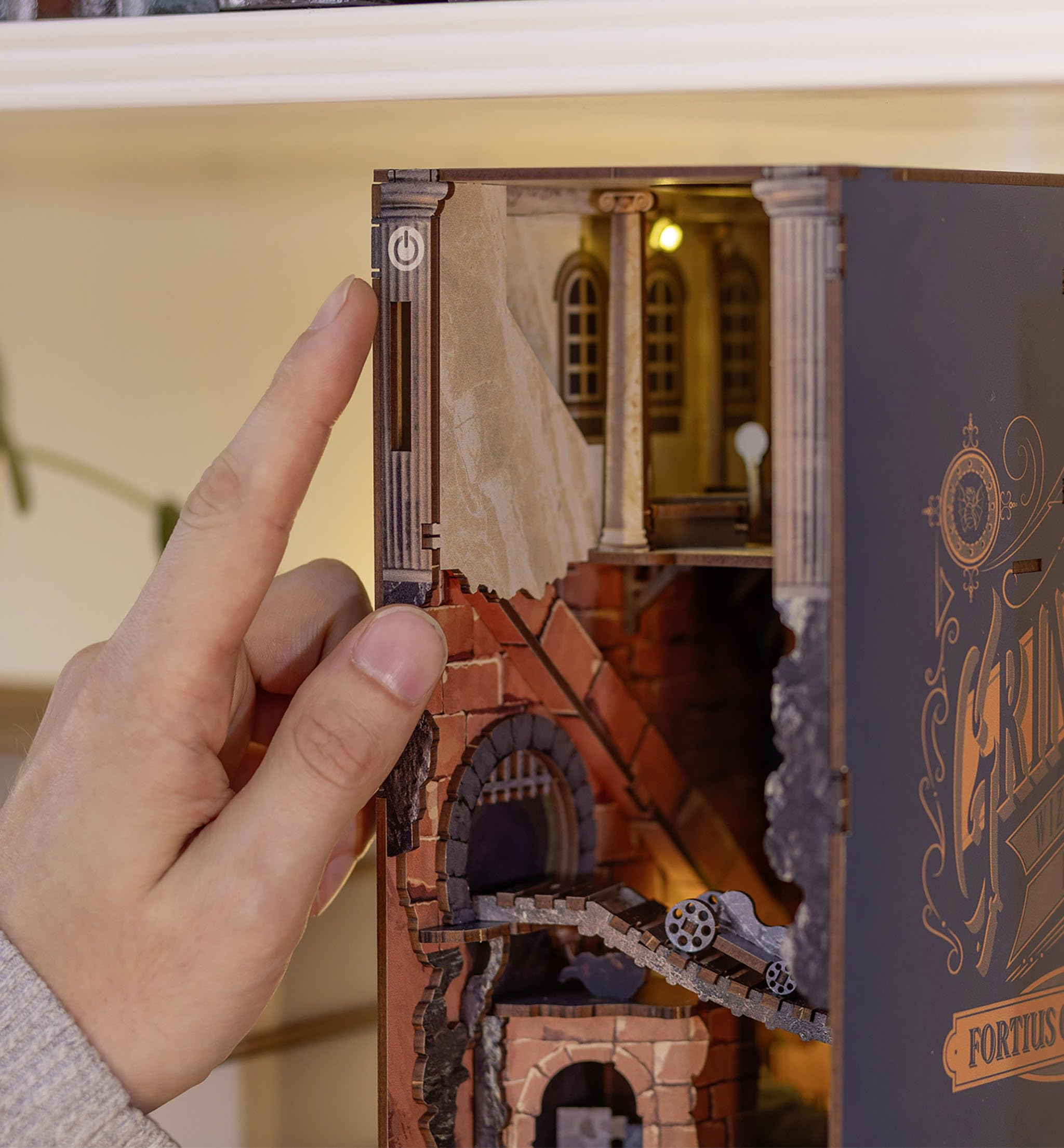 Eine Hand drückt den Power-Knopf eines beleuchteten Holzdioramas mit architektonischen Details.