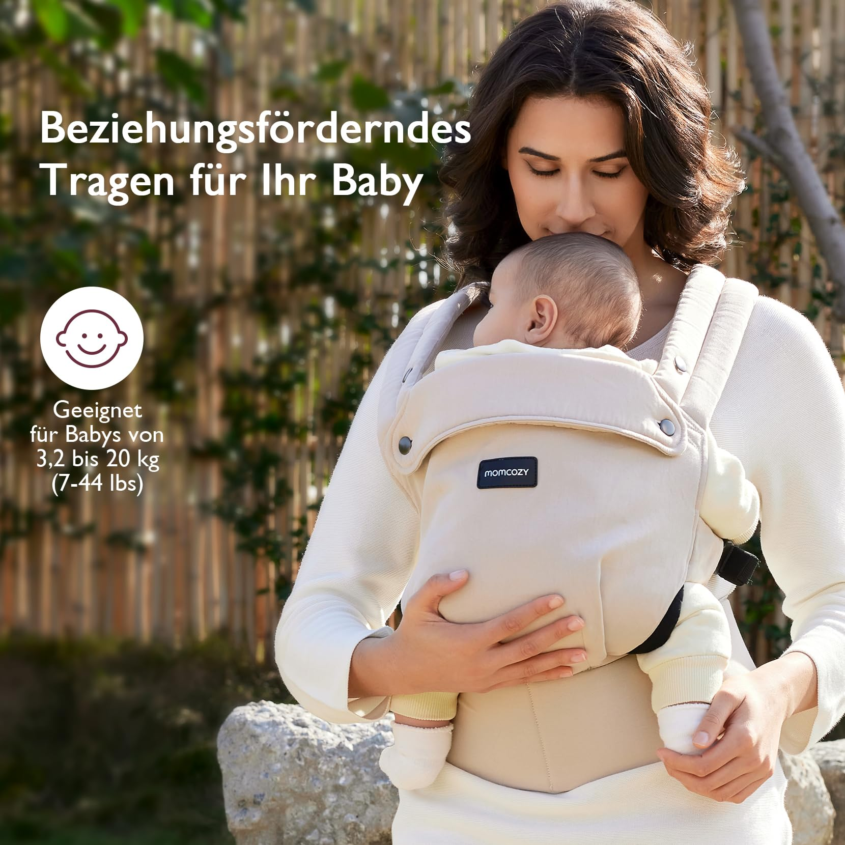 Eine Frau trägt ein Baby in einer cremefarbenen Babytrage vor einem Bambushintergrund.