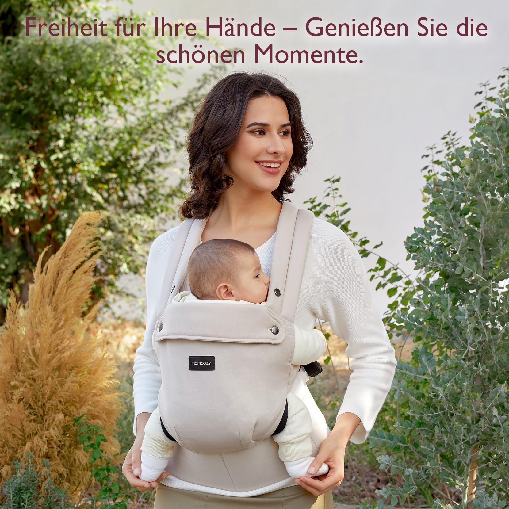 Eine Frau trägt ein Baby in einer cremefarbenen Babytrage in einem Garten.