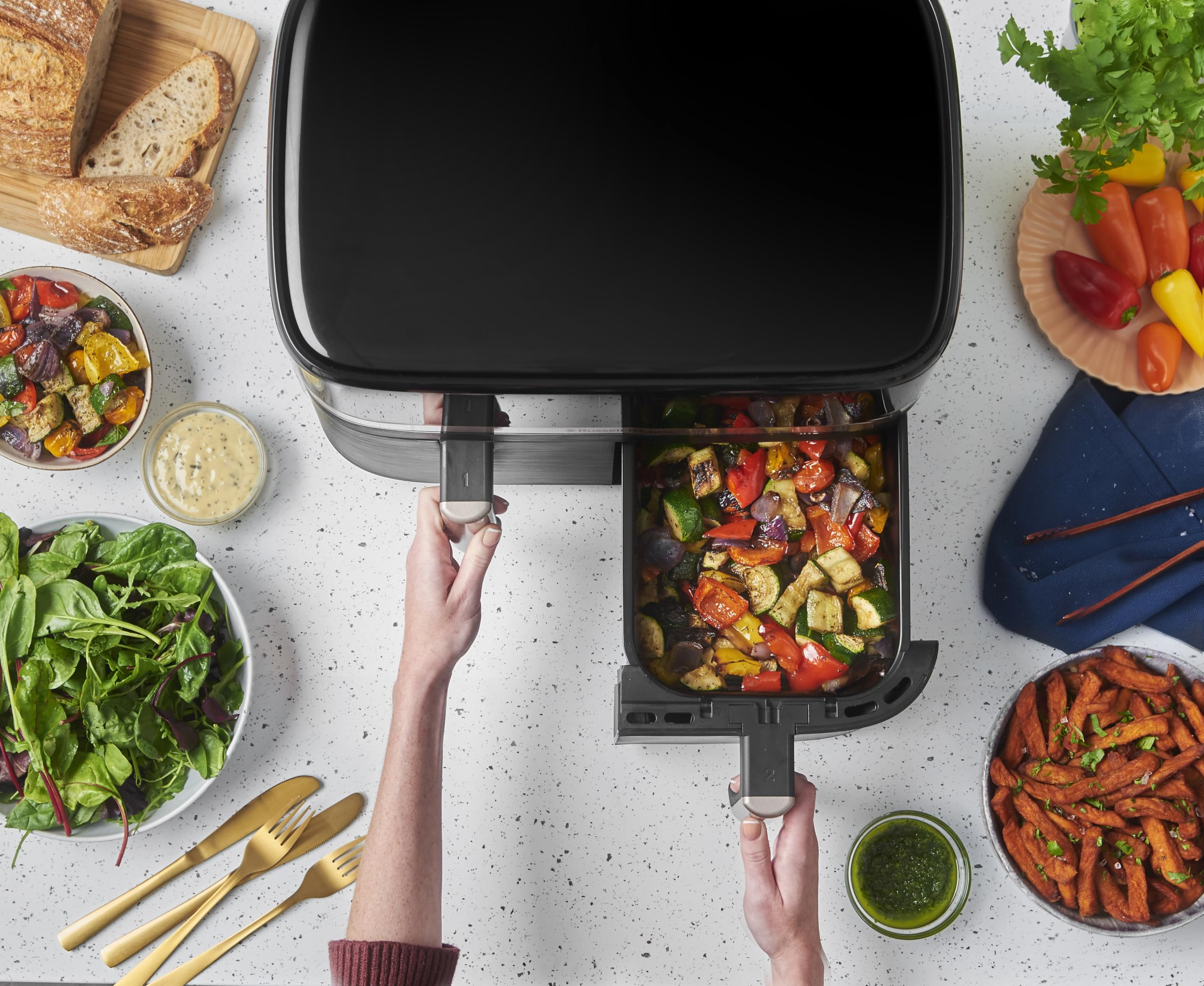 Vue plongeante d'une friteuse à air ouverte contenant des légumes rôtis, entourée de divers aliments.