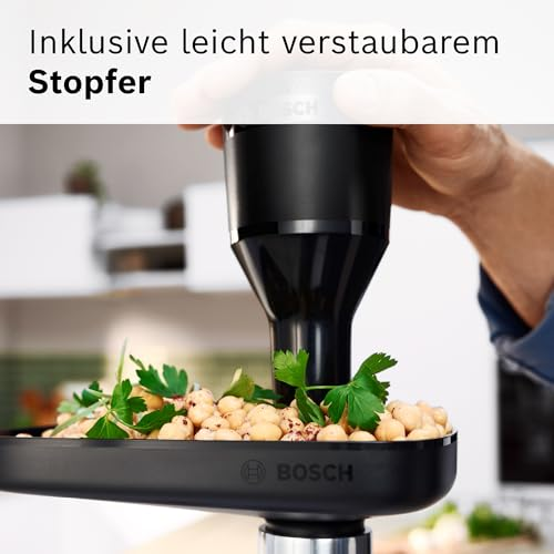 Eine Hand benutzt einen schwarzen Stopfer auf einer Bosch-Küchenmaschine, um Kichererbsen und Kräuter zu drücken.